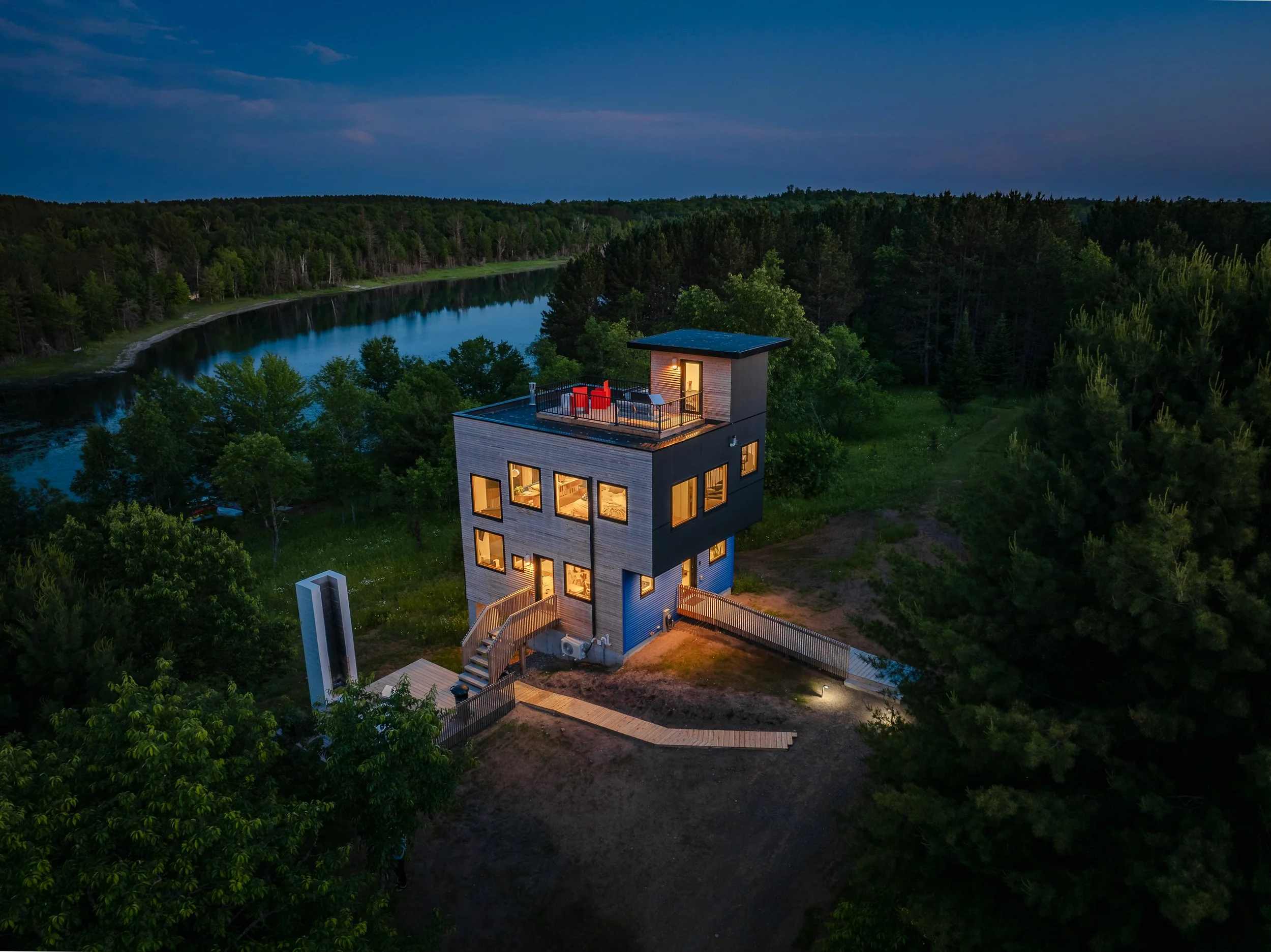 A modern multi-story house with illuminated windows and a rooftop terrace surrounded by trees, overlooking a river at dusk.