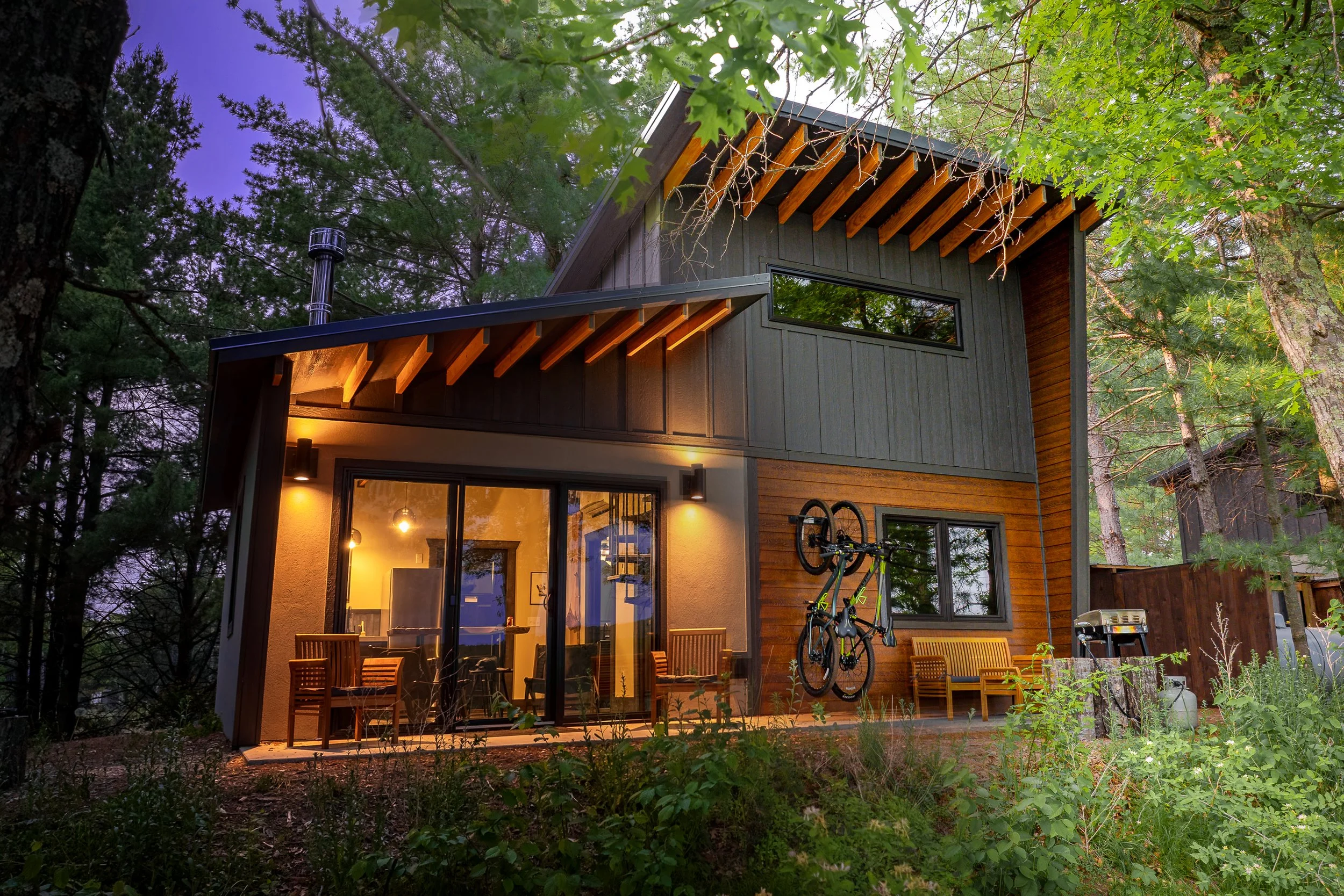 Modern two-story house in a wooded area with large glass sliding doors, wooden benches, a bicycle mounted on the wall, and an outdoor grill, illuminated at dusk.