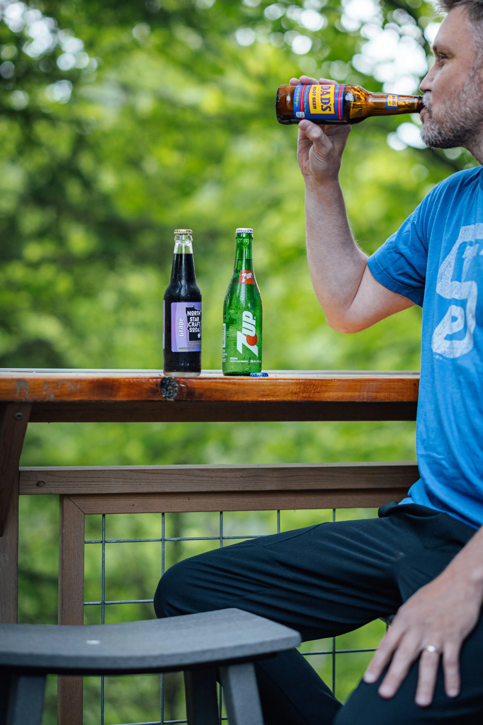 A man with gray hair and beard sitting at an outdoor wooden table, drinking from a brown glass bottle of Daris beer, with two other bottles, one labeled North Star Craft Soda and the other labeled 7Up, on the table. The background shows green trees a
