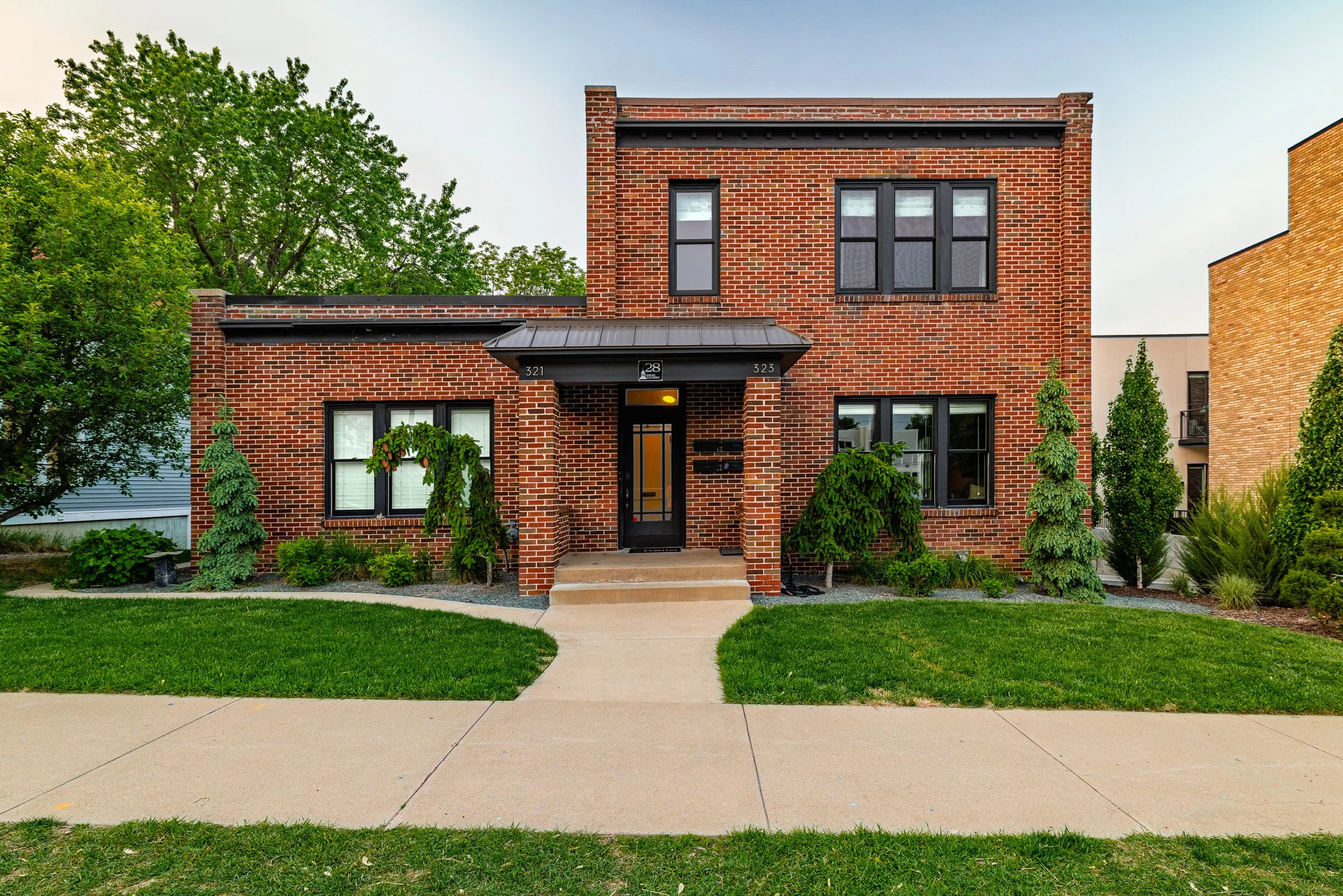 A brick residential building with black window frames and a black front door, surrounded by a lawn and landscaped bushes, trees, and a concrete sidewalk walkway.