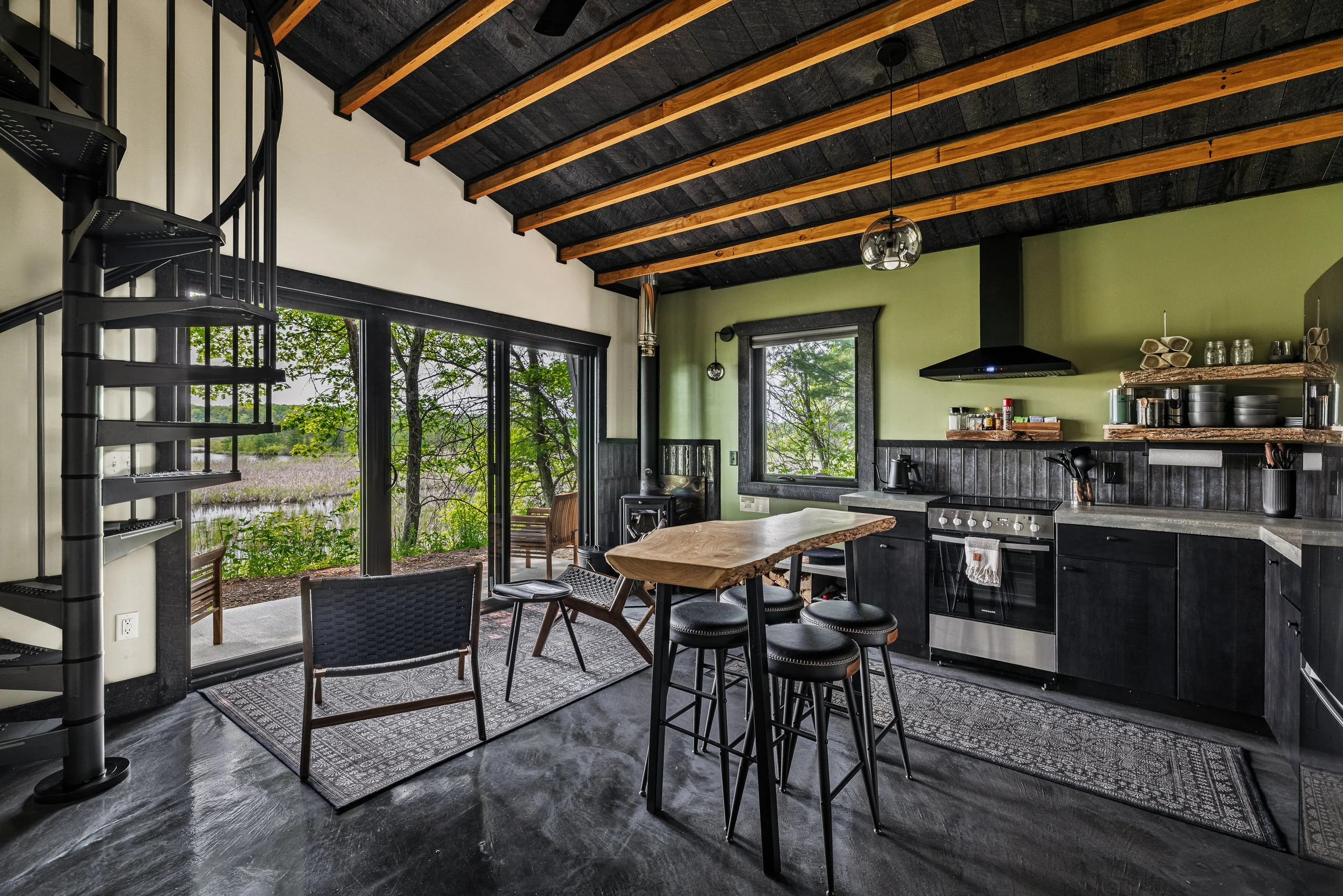 Kitchen with black cabinets, a wooden table with four stools, a black spiral staircase, large sliding glass door opening to a porch with outdoor seating, green walls, and views of trees outside.