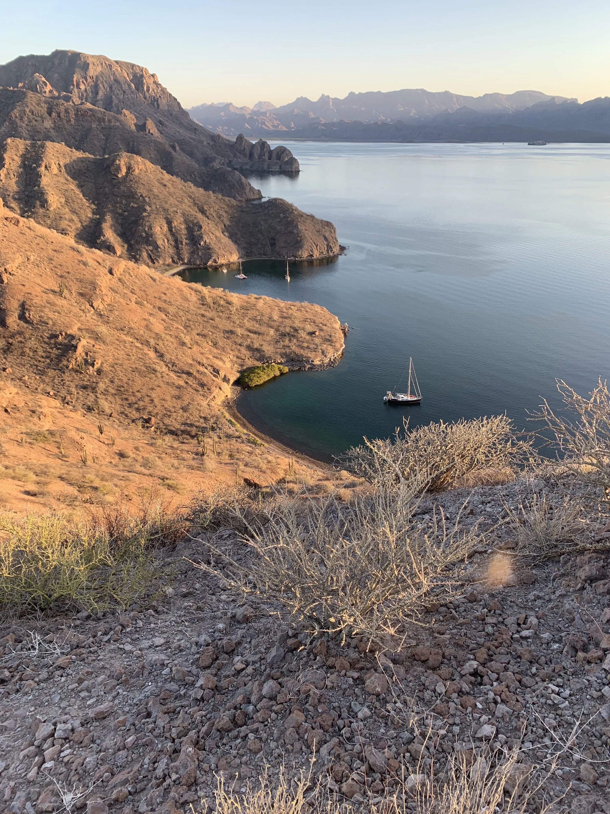 Scenic view of rugged desert mountains and a calm lake with sailboats at sunrise or sunset.
