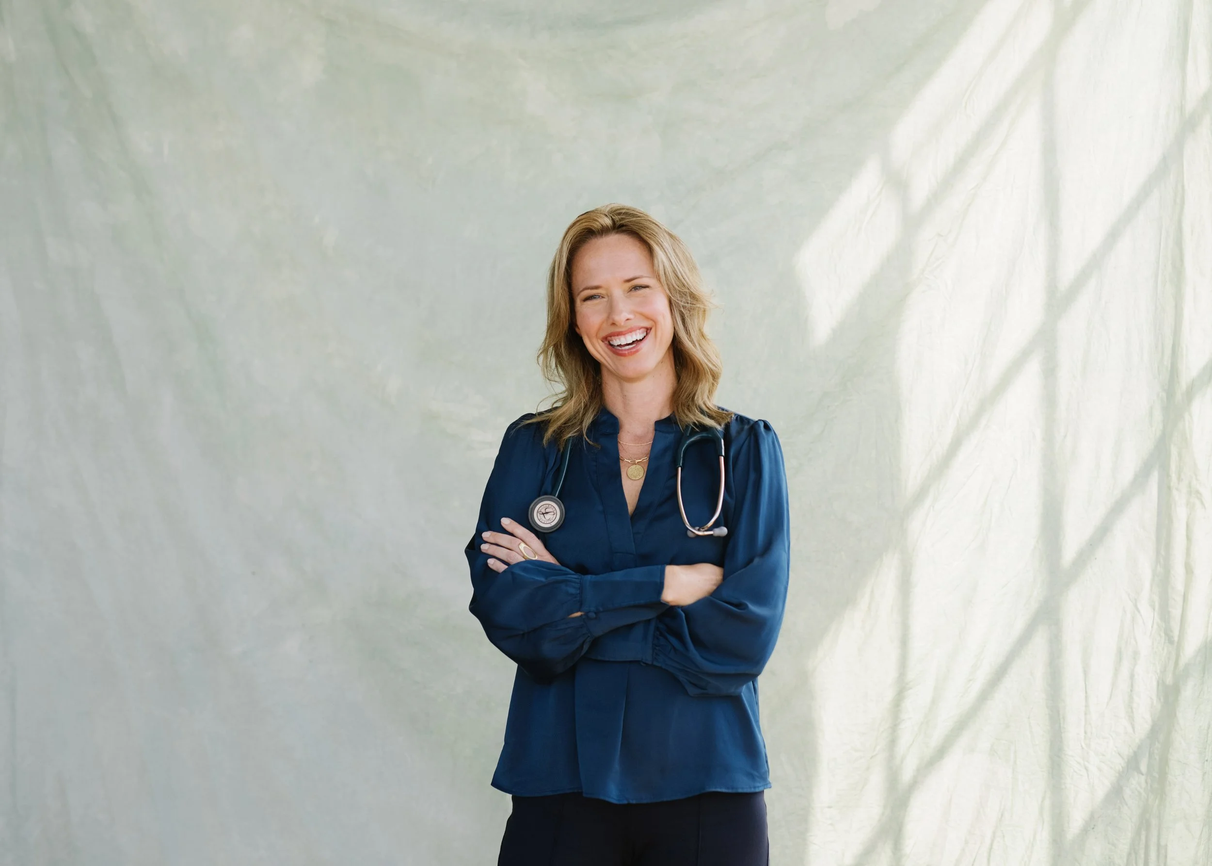 A smiling female doctor with a stethoscope around her neck standing with arms crossed in front of a light-colored, textured background.