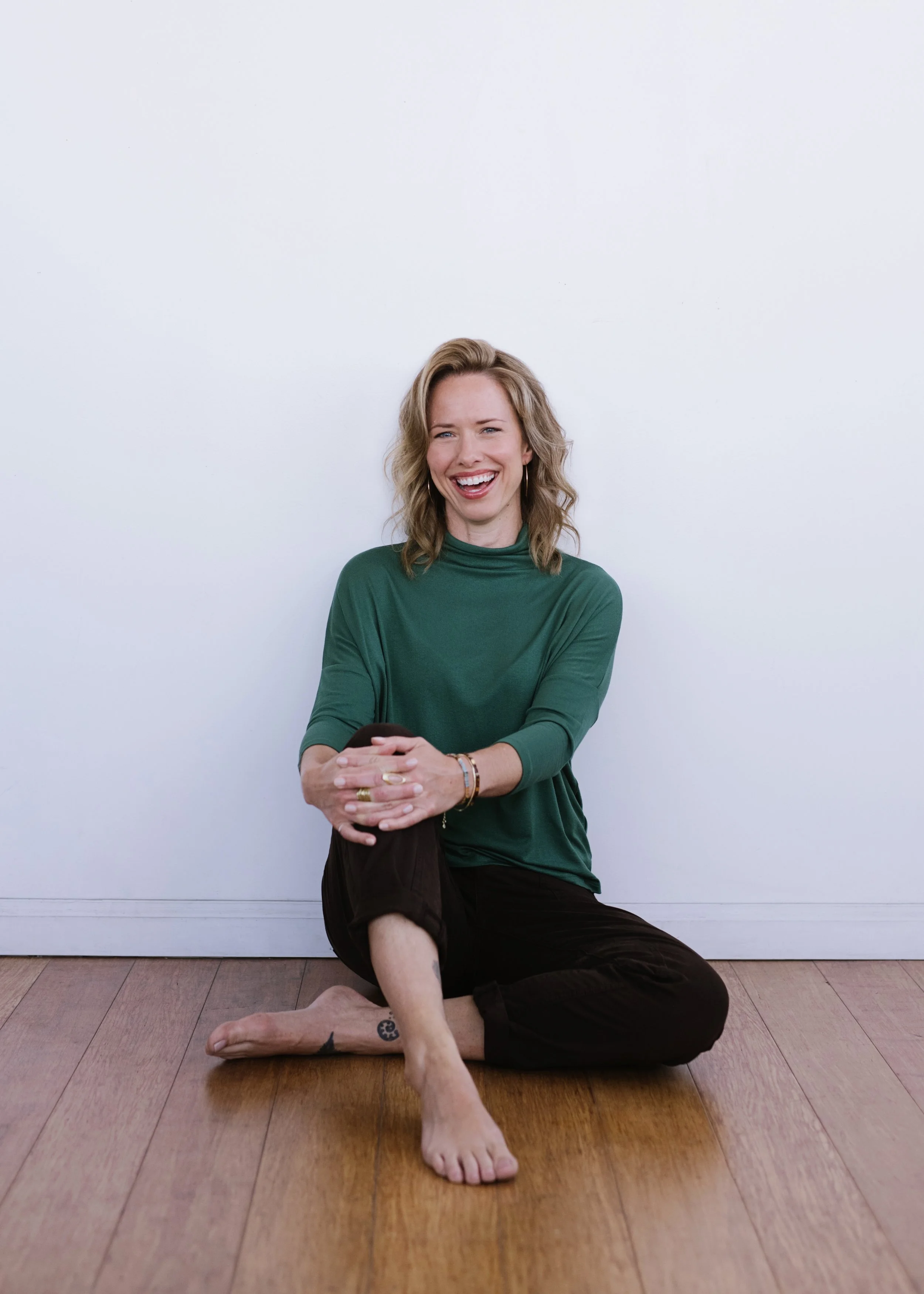 A woman with wavy blonde hair, wearing a green long-sleeve top and black pants, sitting on a wooden floor against a plain white wall, smiling and laughing while holding a phone in her hands.