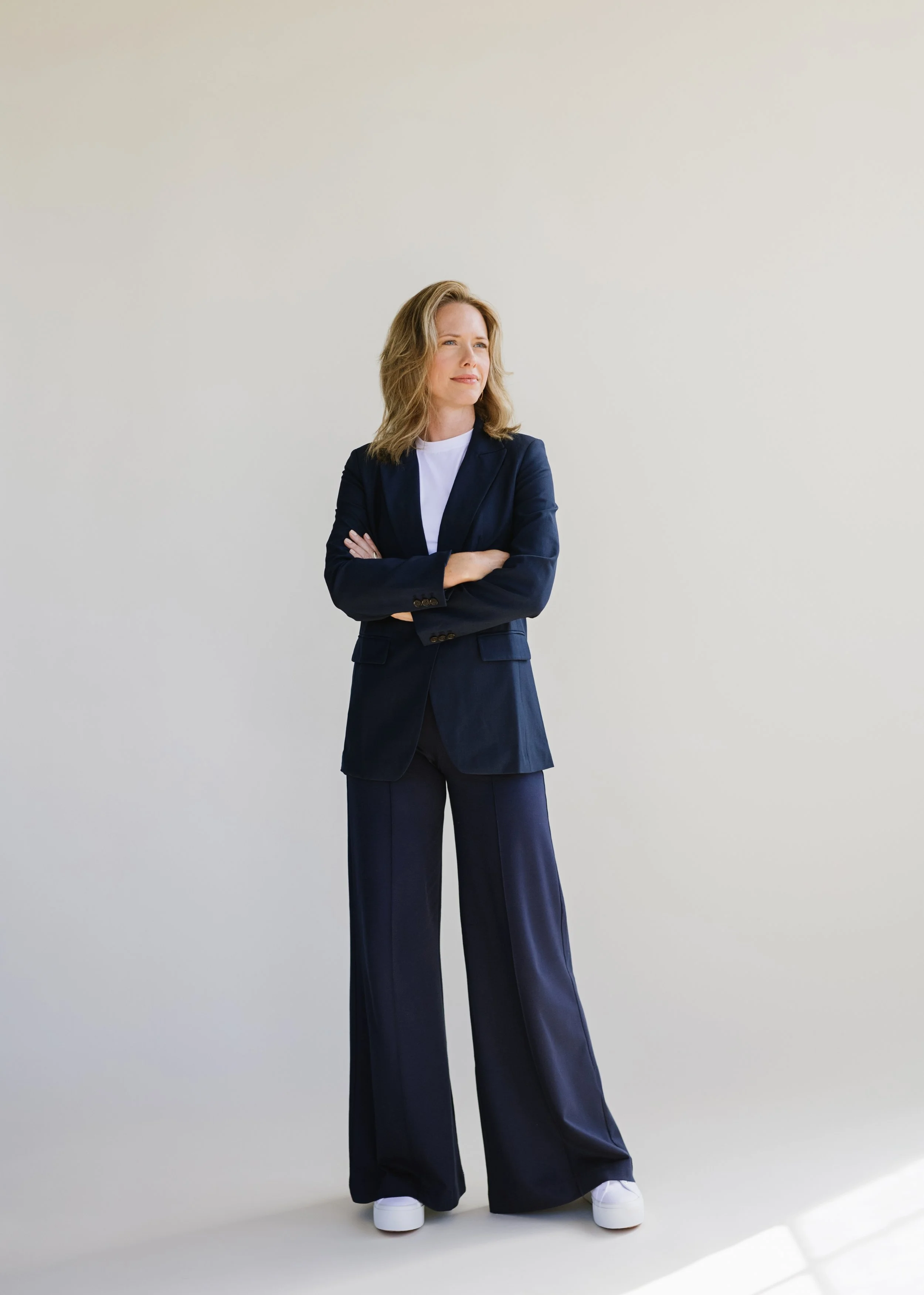 A woman with shoulder-length blonde hair dressed in a navy blazer, matching wide-leg pants, white shirt, and white sneakers, standing against a plain off-white background with her arms crossed, looking slightly to the side.