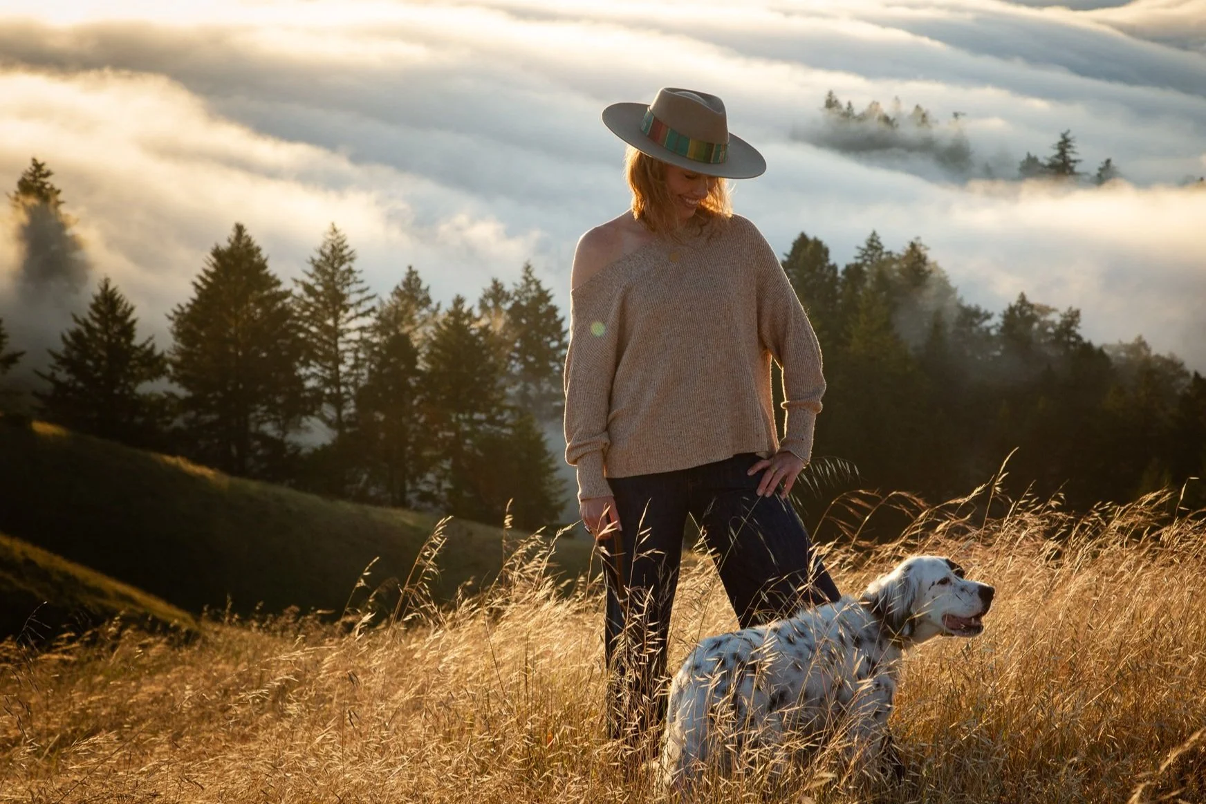 A woman in a beige sweater and wide-brimmed hat standing in a grassy field with a white dog during sunset, with a forest and fog in the background.