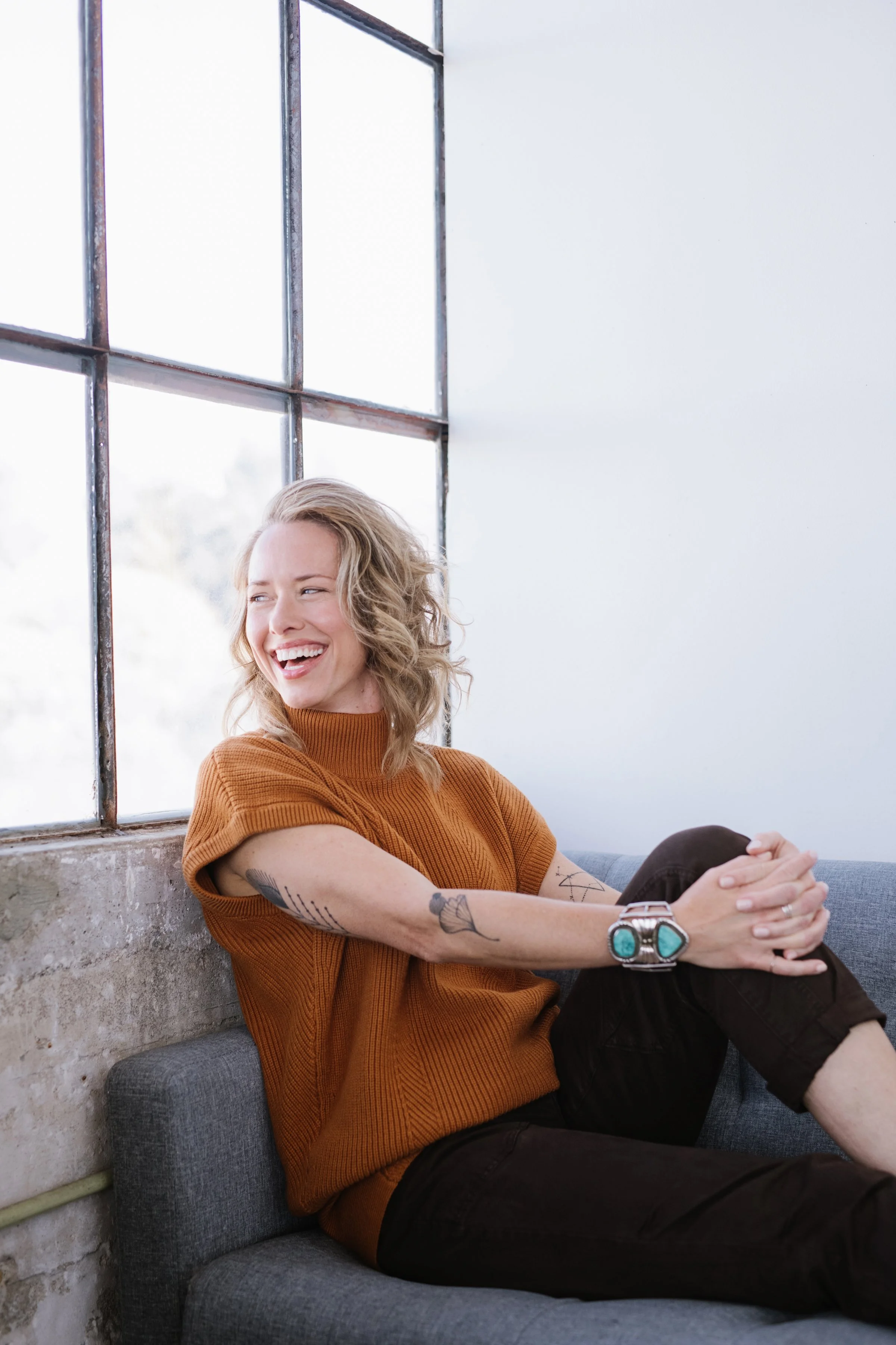 A woman with blonde hair, tattoos on her arm, wearing an orange sweater and black pants, sitting on a gray couch near a large industrial-style window, smiling and looking to her left.