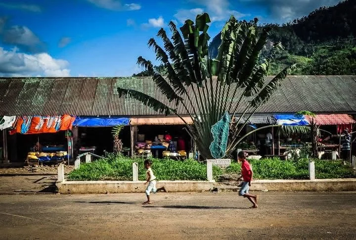 Two children running on the street with market stalls and a large banana tree in the background, under a blue sky with some clouds, in a rural area.