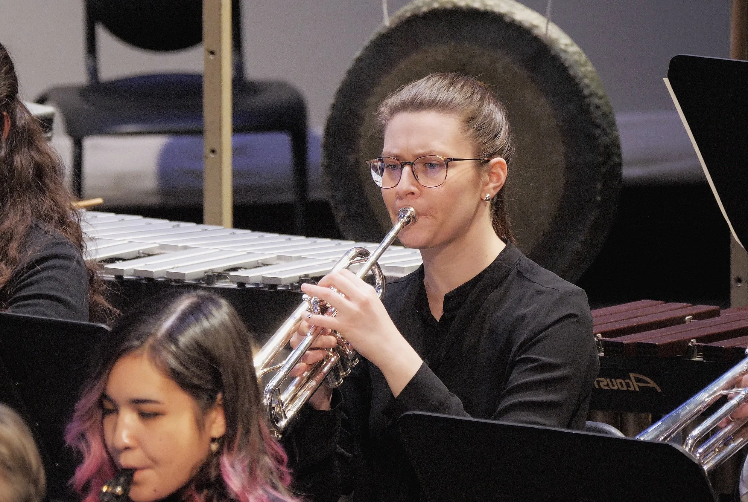 A woman with glasses and a black shirt playing the trumpet in an orchestra, with a large gong and a xylophone in the background.