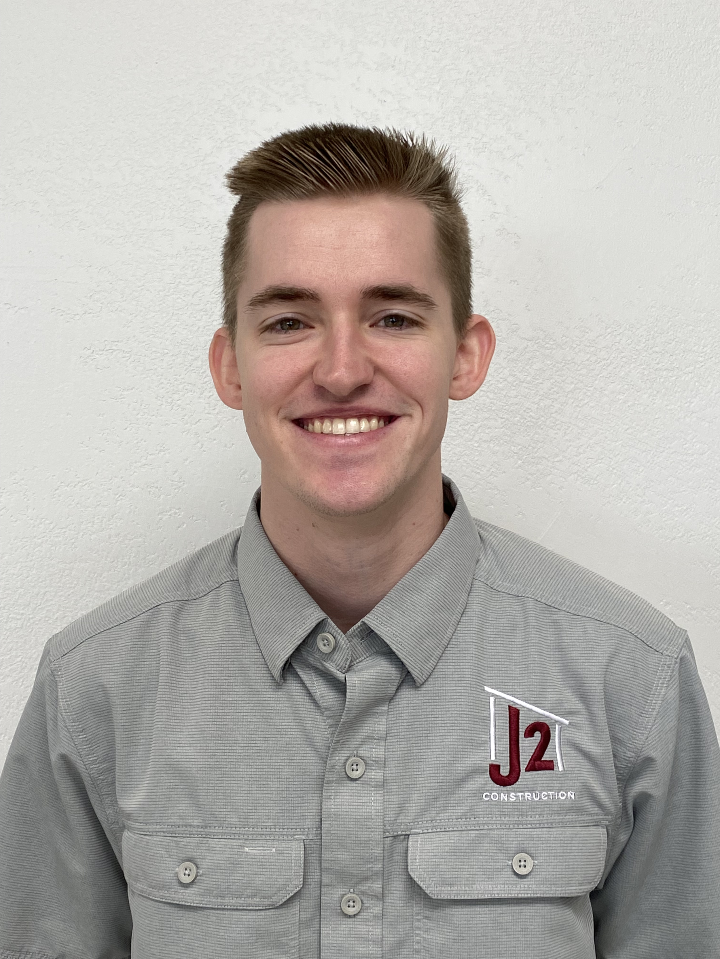 Young man wearing a light gray uniform shirt with the logo 'J2 Construction' on the chest, smiling in front of a plain white wall