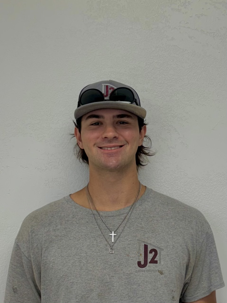Young man smiling, wearing a gray T-shirt with 'J2' logo, a baseball cap, sunglasses on the cap, and necklaces with crosses, standing against a white wall.