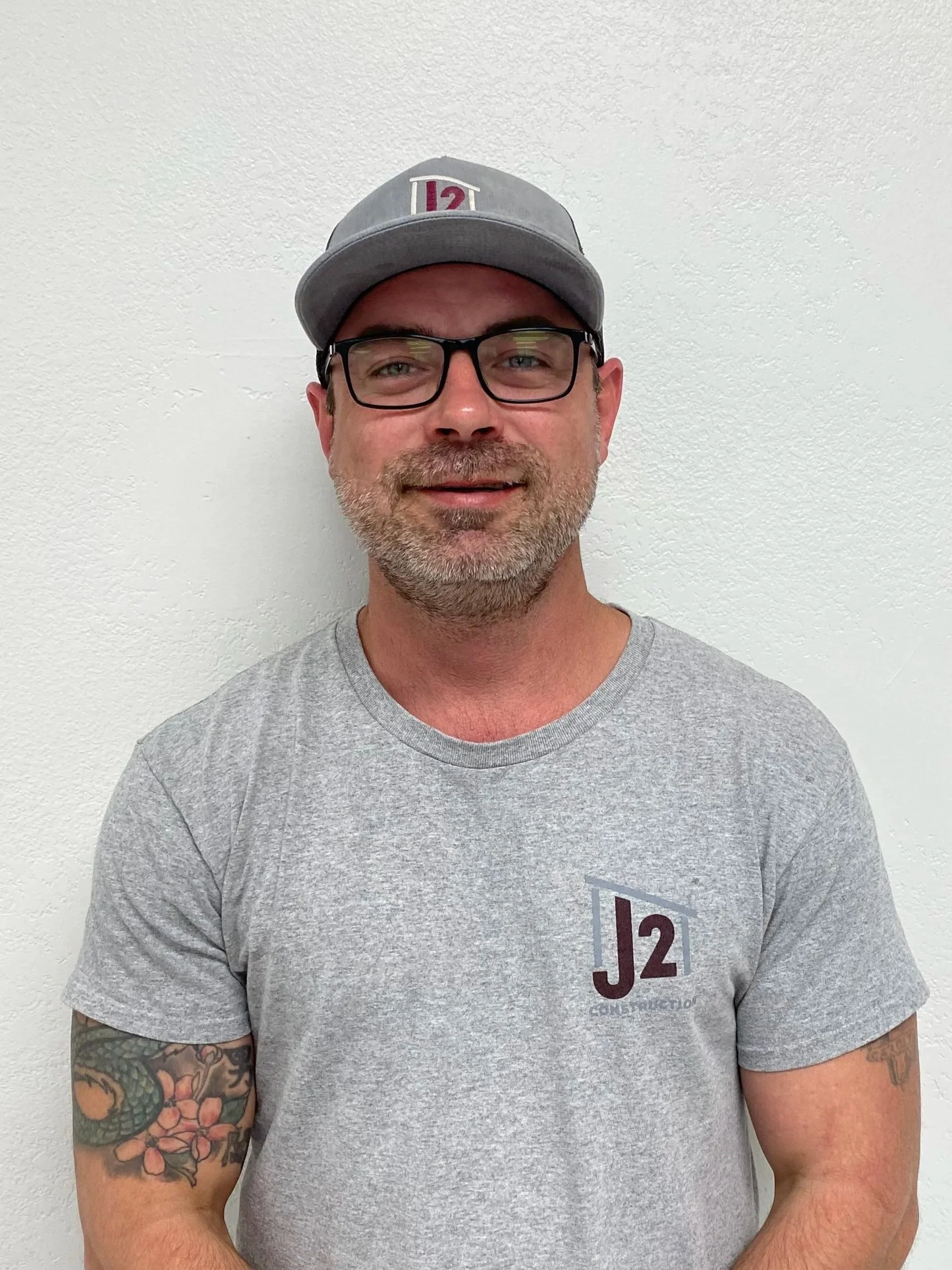 A man with glasses and a gray baseball cap with the number '12' on it, wearing a light gray T-shirt with a logo, standing against a white textured wall.