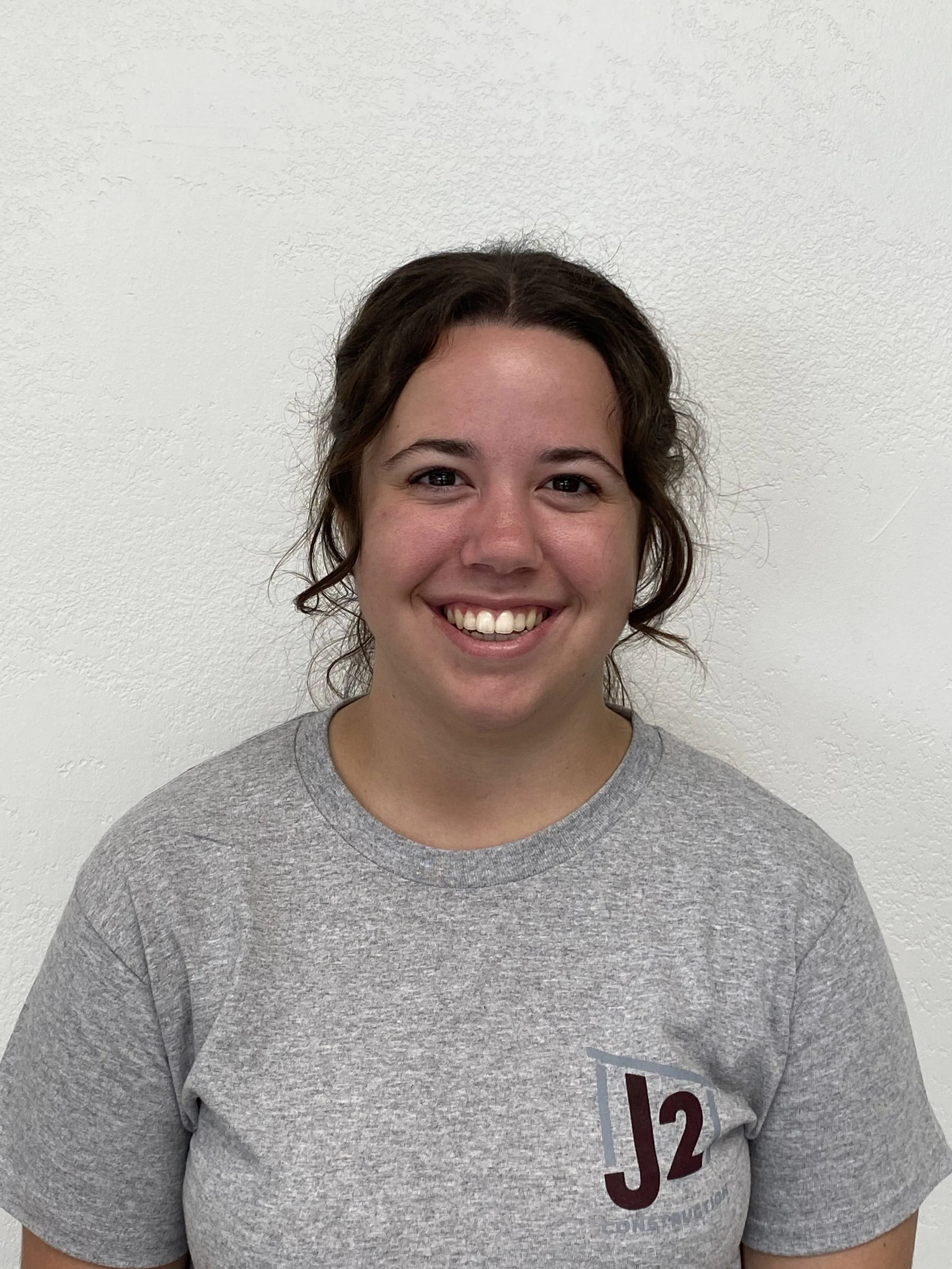 A young woman smiling with braces, wearing a gray t-shirt, in front of a plain white wall.