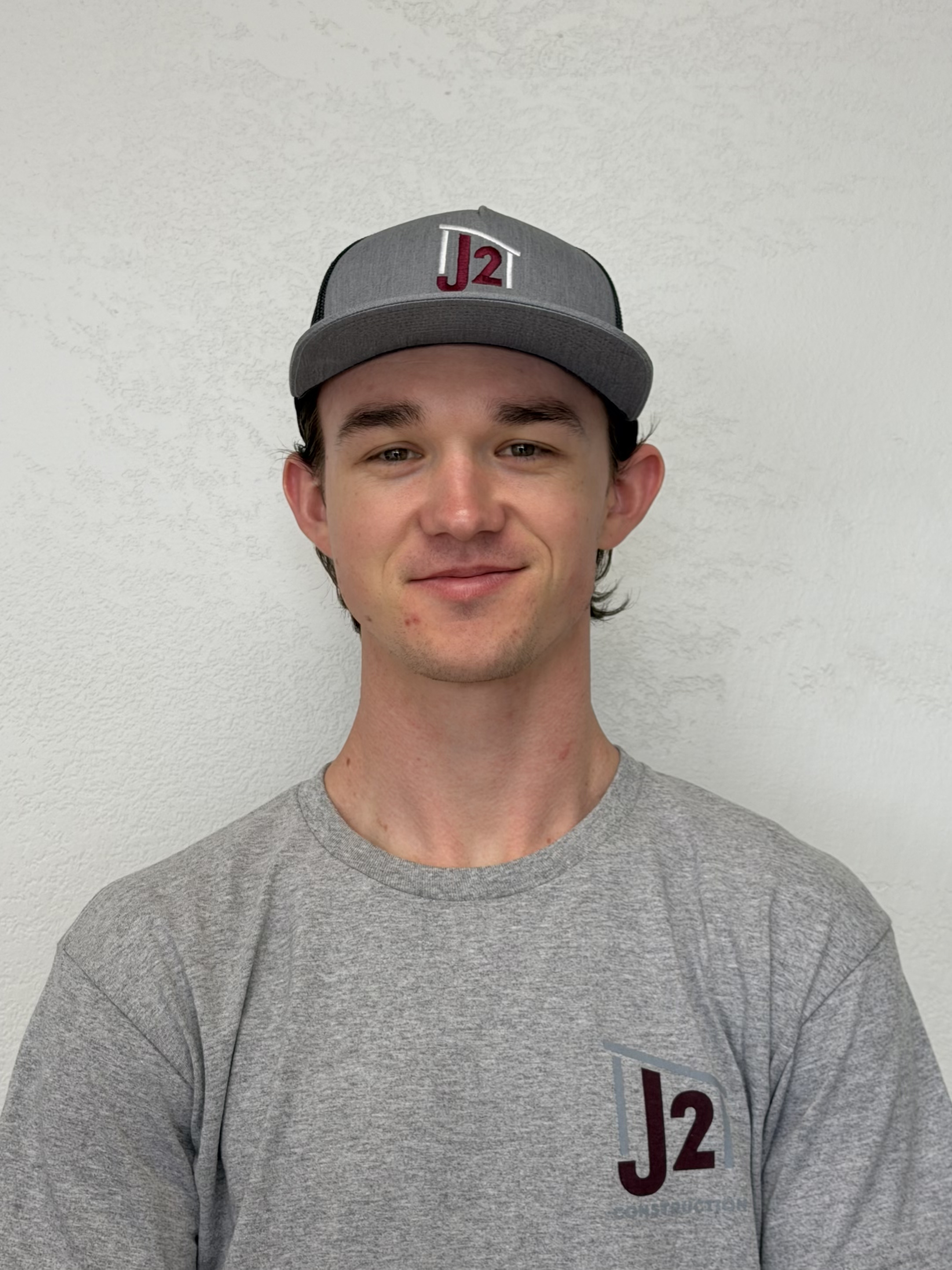 A young man with dark hair wearing a gray baseball cap and a gray T-shirt, standing against a white textured wall.