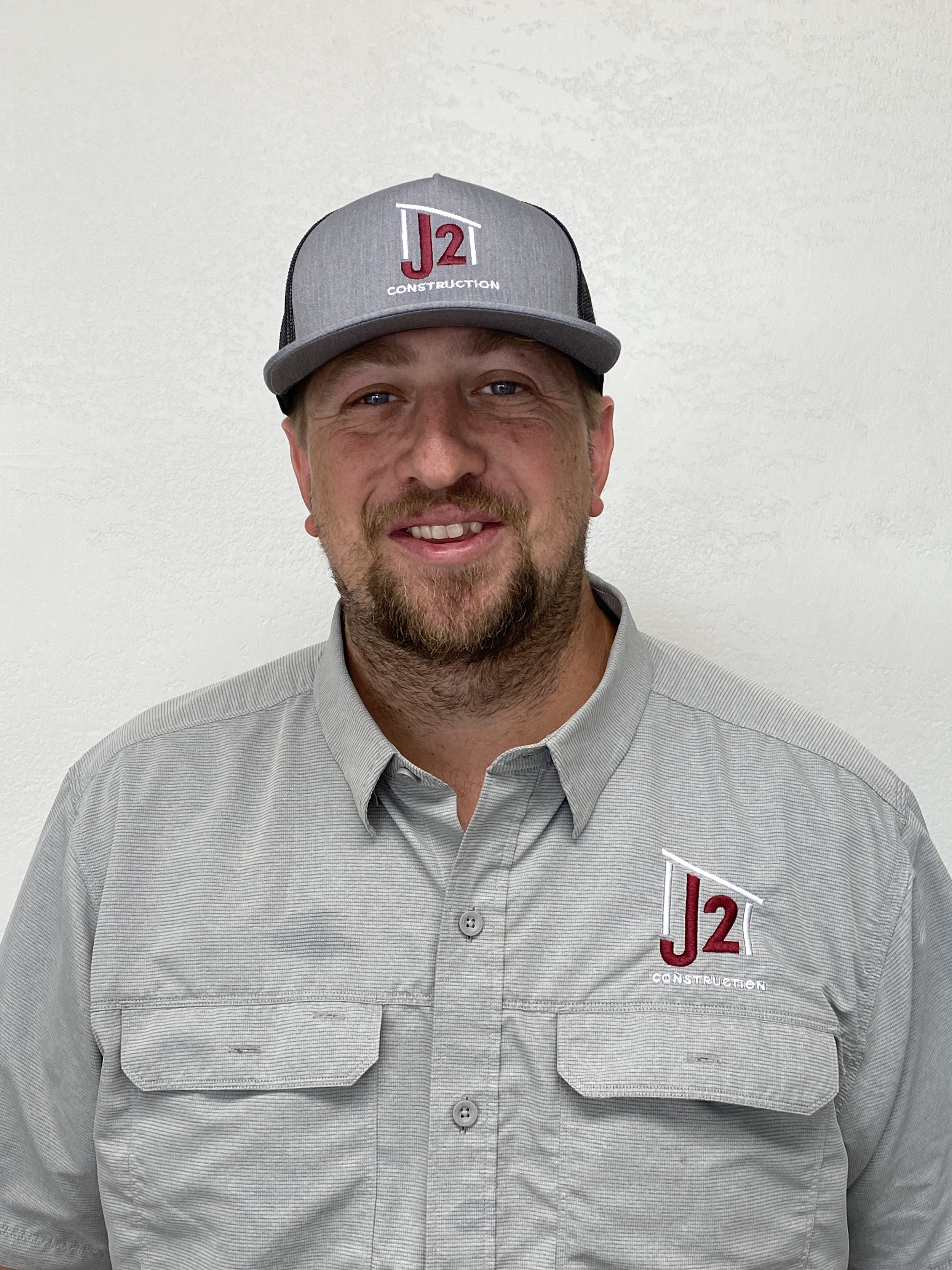 A man wearing a gray cap with a logo that says 'J2 Construction' and a light gray button-up shirt with the same logo on the left side. The background is plain white.