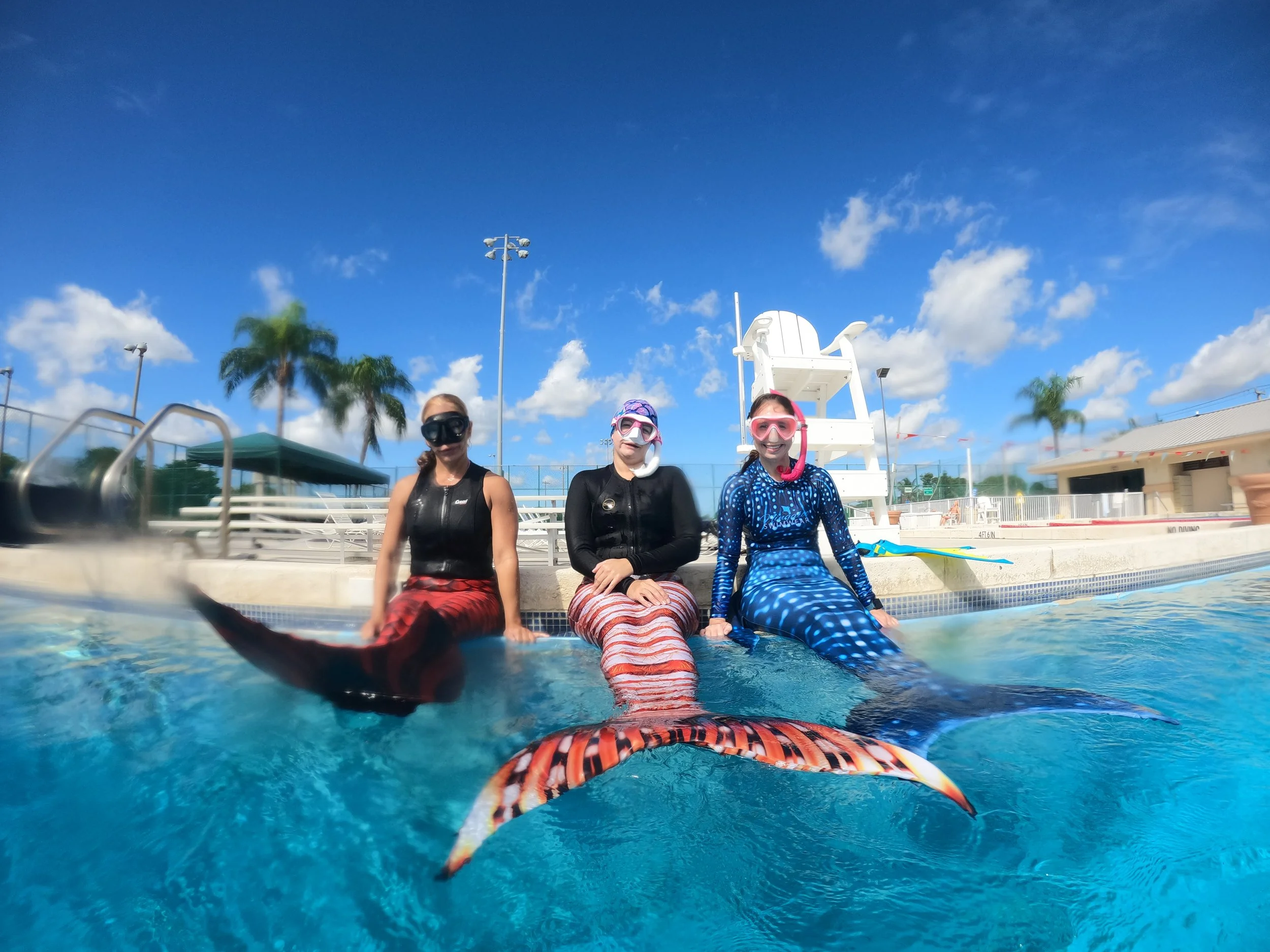 Three women wearing mermaid tails and snorkeling gear sitting on the edge of a swimming pool with a blue sky, palm trees, and poolside structures in the background.