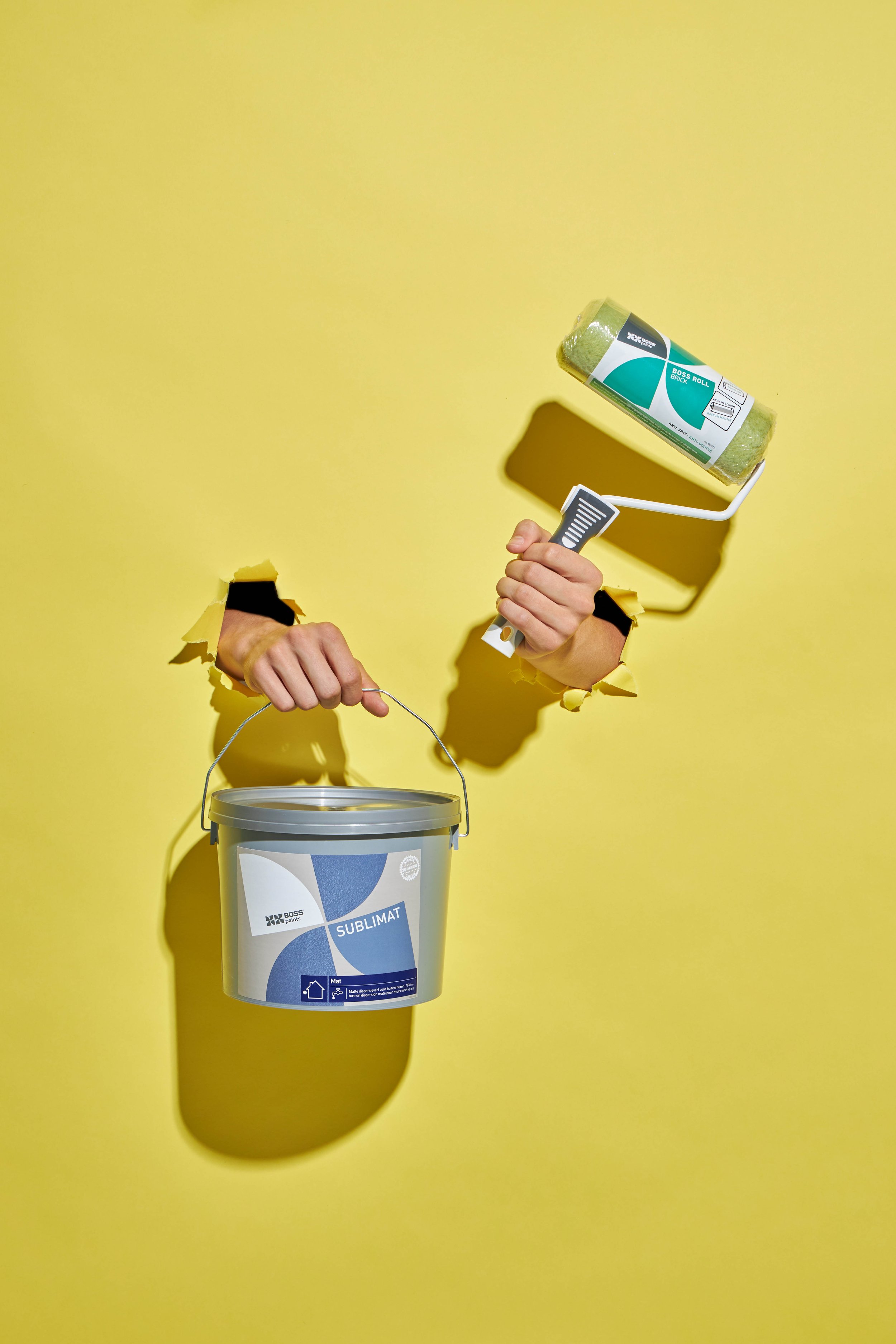 Productfoto voor BOSS Paints gemaakt in de studio van 2 handen die door de papieren achtergrond komen, één hand heeft een verfpot vast de andere een verfrol van BOSS Paints, gefotografeerd door Simon van Rhee
