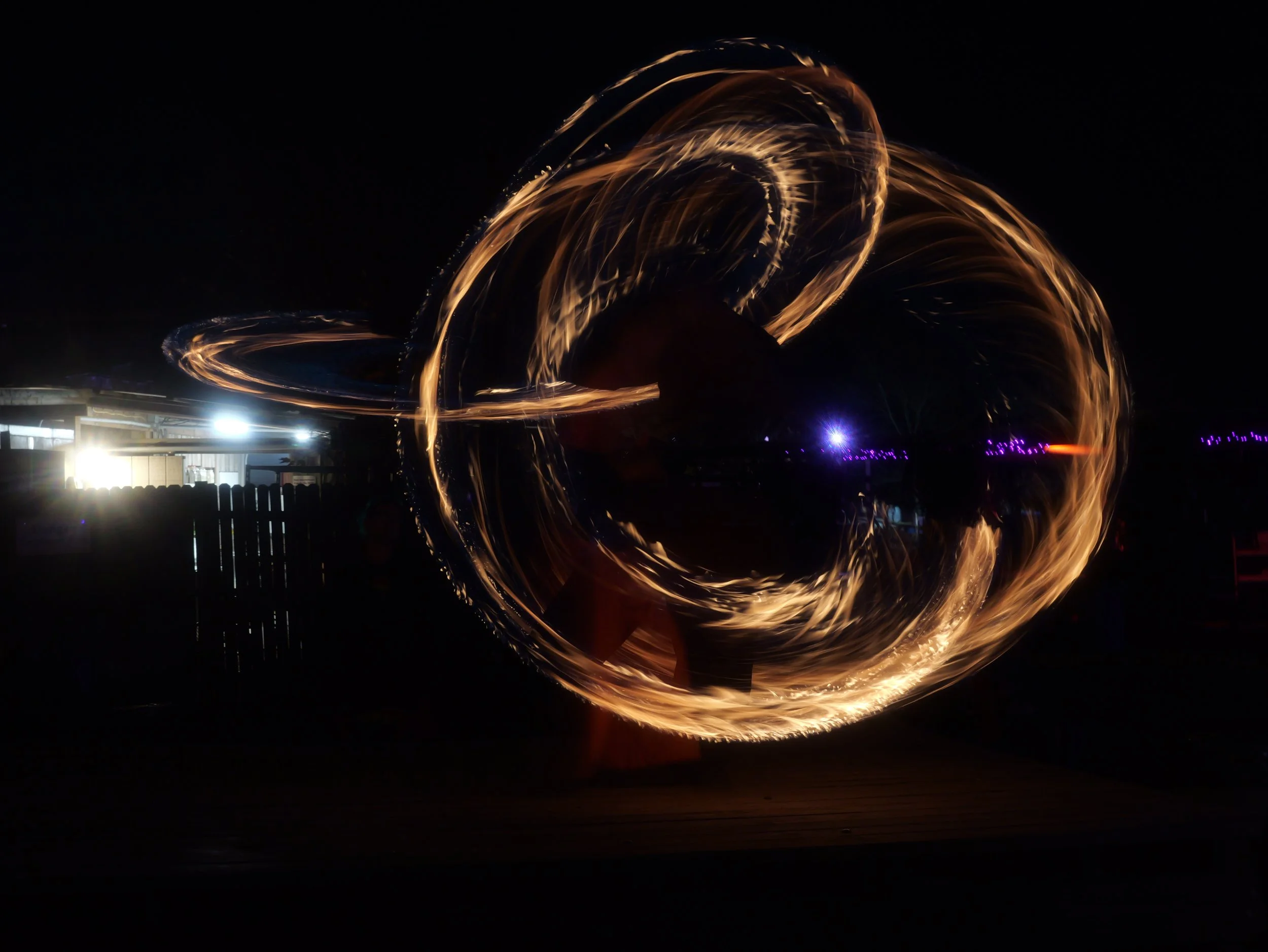 Long exposure photo of a person performing steel wool spinning at night, creating bright fiery trails in circular patterns against a dark background.