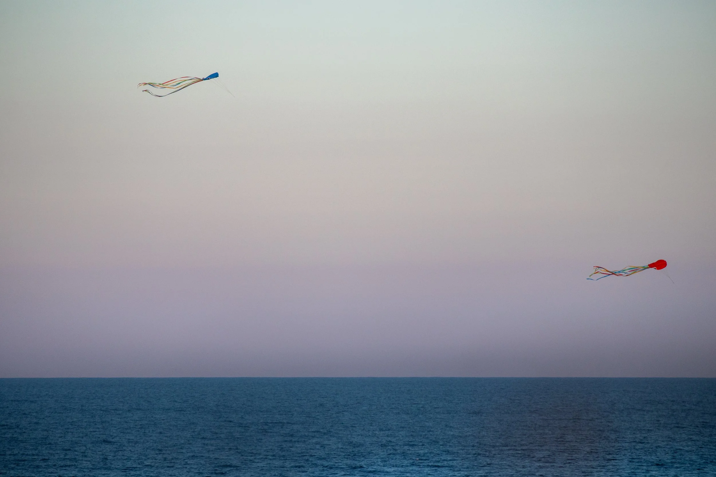 Two colorful plastic wings with long strips of fabric attached, flying over the ocean on a cloudy day.