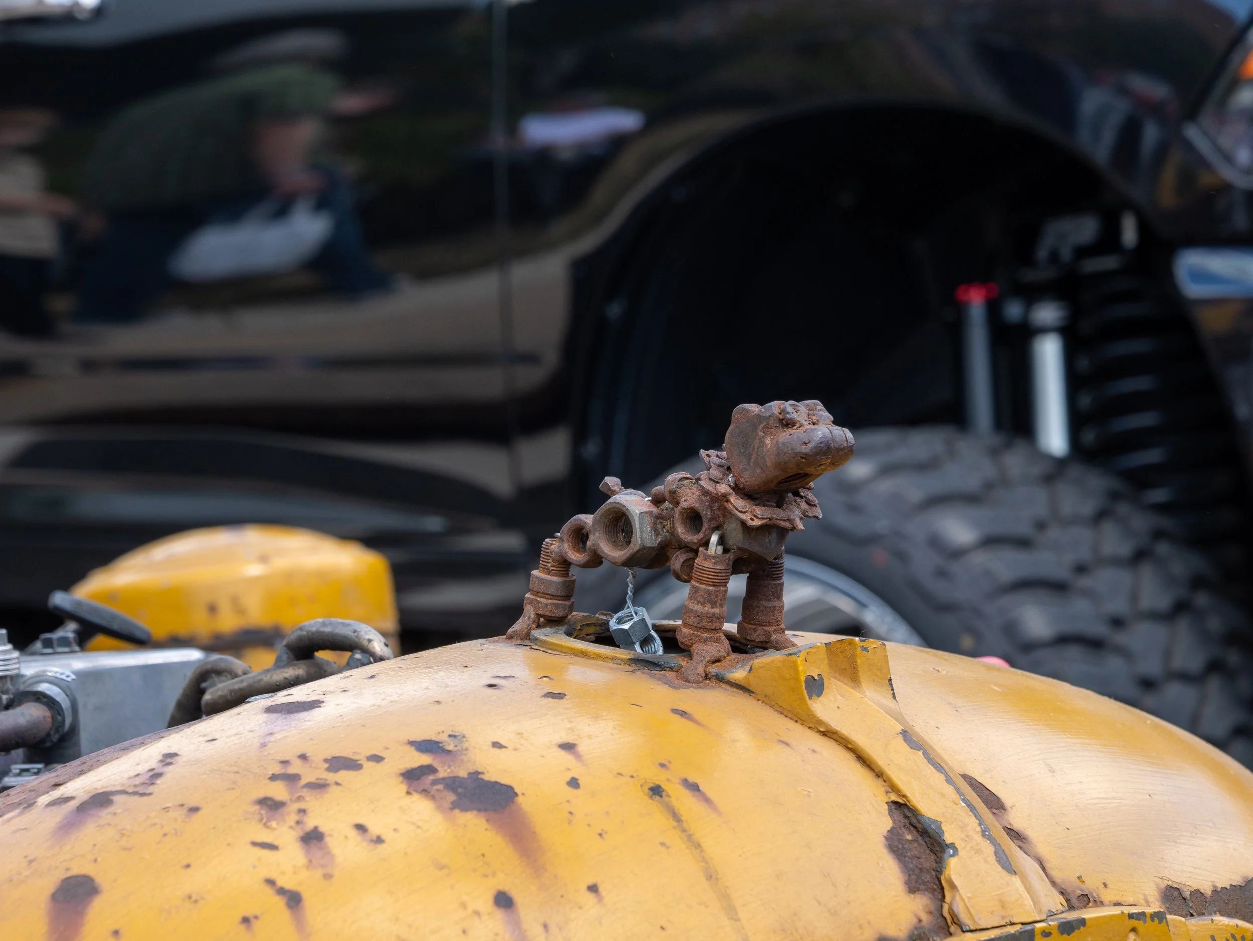 Rusty mechanical part mounted on a yellow surface, with a black automobile and a person reflected in the background.