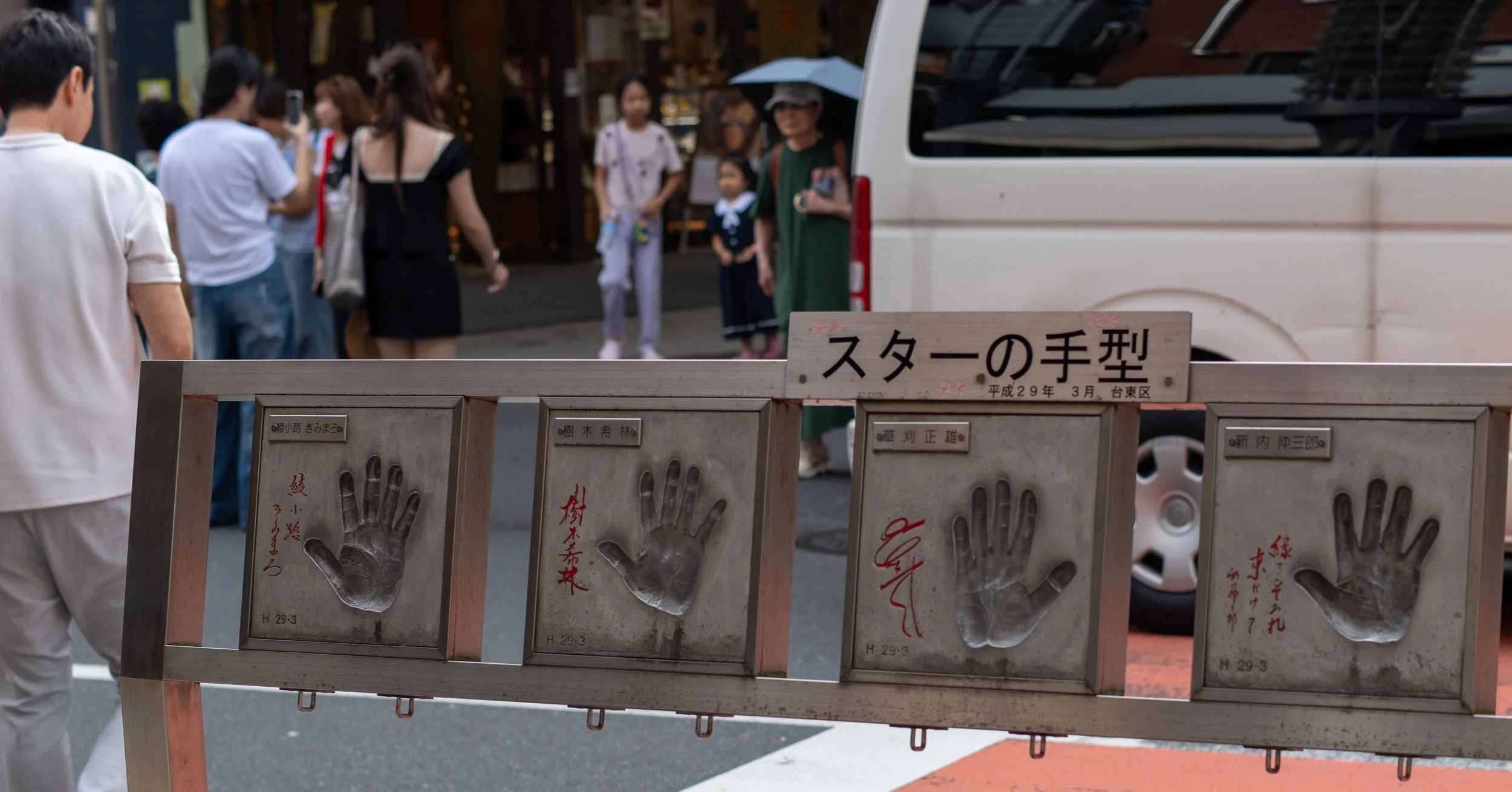 A display with multiple handprints and signatures on wooden panels in front of a busy street with several people, some with umbrellas, and vehicles parked along the street.