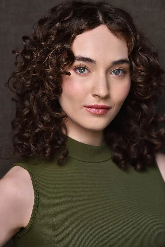 Theatrical headshot of an actor with curly brown hair and green eyes wearing an olive-green sleeveless top against a dark brown backdrop.