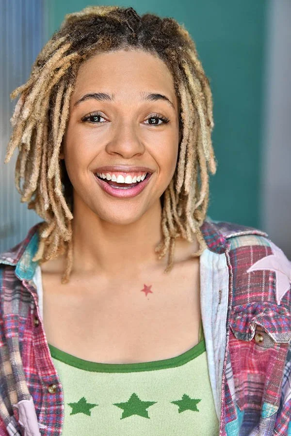 Expressive commercial headshot of a smiling young Black woman with blonde-tipped locs wearing a plaid shirt and green star top.