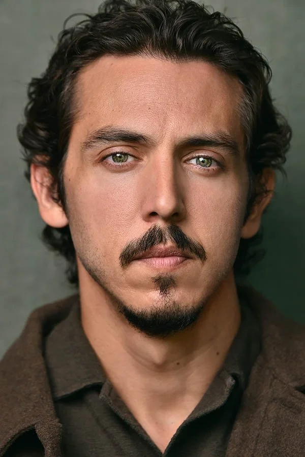 Intense dramatic actor headshot featuring a dark-haired male actor with a goatee and facial hair, wearing an earthy brown jacket against a neutral studio backdrop. Captured by Los Angeles headshot photographer Marc Cartwright.