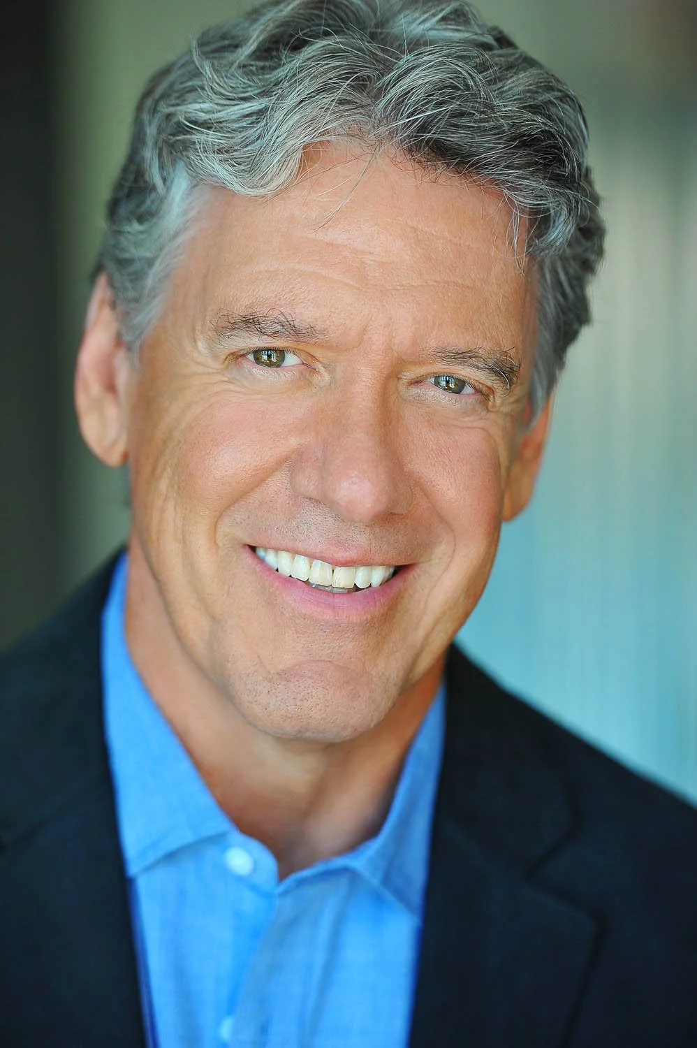 Actor headshot of a gray-haired man in a navy jacket and light blue shirt, smiling warmly