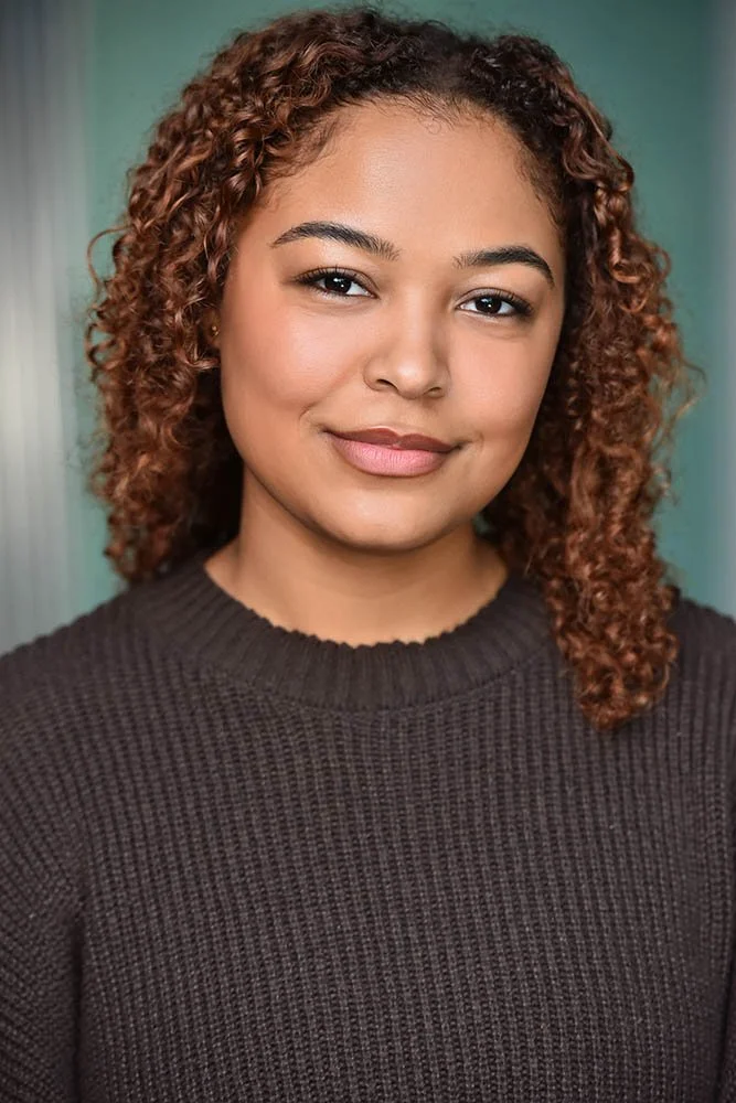 Actor headshot of a woman with long auburn curls in a brown knit sweater, smiling softly against a teal background