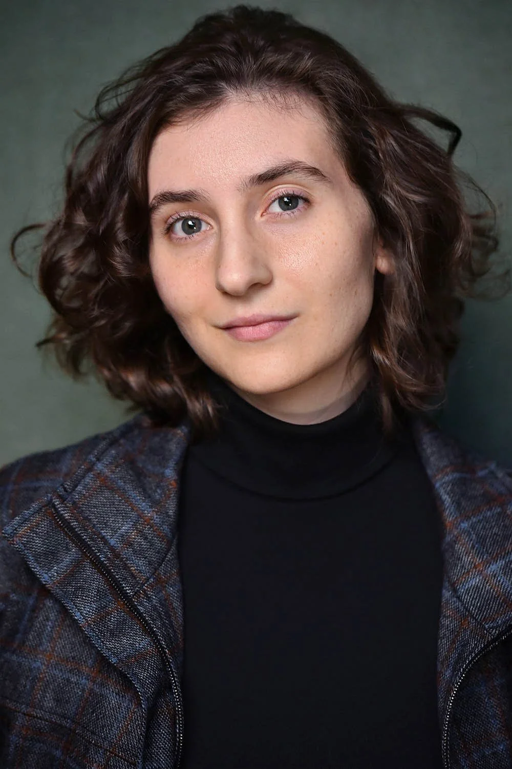 Headshot of a woman with wavy shoulder-length brown hair and a slight smile, wearing a black turtleneck and plaid jacket against a dark green background.