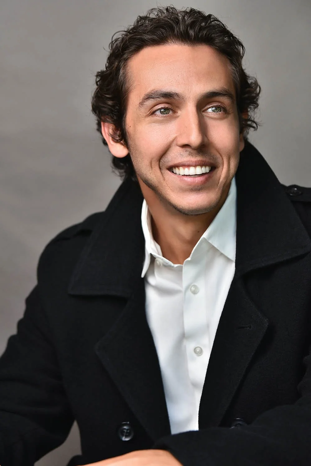 Man with dark wavy hair smiling broadly and looking off to the side, wearing a black wool coat over a white collared shirt against a gray background.