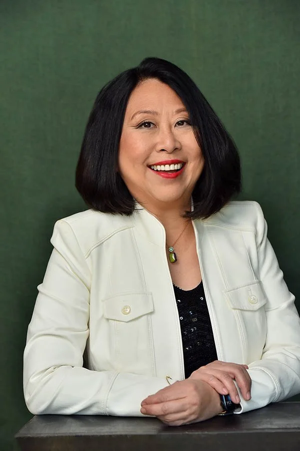 Professional business woman seated at a table, smiling confidently, wearing a light-colored jacket over a dark top, hands resting naturally on the tabletop against a muted green studio background. Photo by Marc Cartwright in Los Angeles