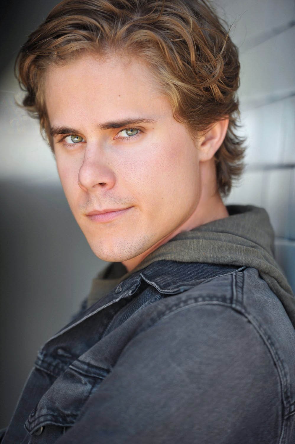 Actor headshot of a man with wavy hair in a gray denim jacket, turned toward the camera against a light painted wall