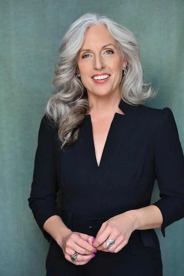 Full-frame professional business headshot of a smiling woman with grey hair in a black dress, showcasing confidence for corporate roles or executive portraits in LA.