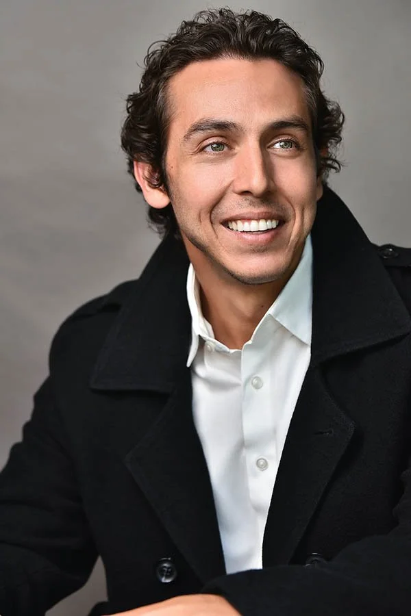 Warm and approachable commercial actor headshot showcasing a smiling man with green eyes, open collar white shirt, and black pea coat against a grey background. Shot by top Los Angeles headshot photographer Marc Cartwright.