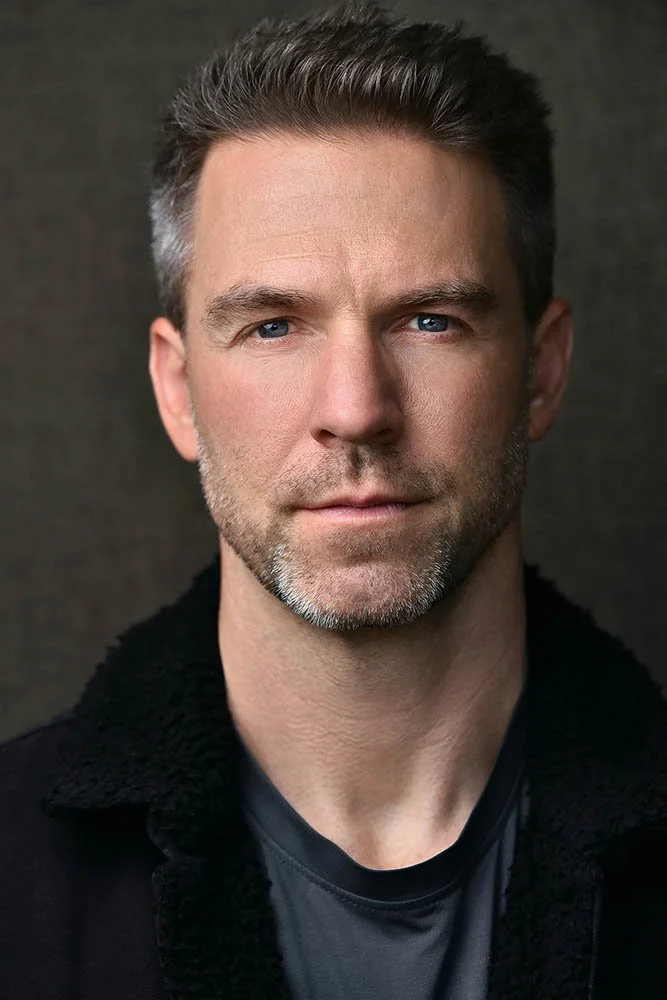 Actor headshot of a man with short graying hair and stubble in a black jacket, looking directly into the camera against a dark background