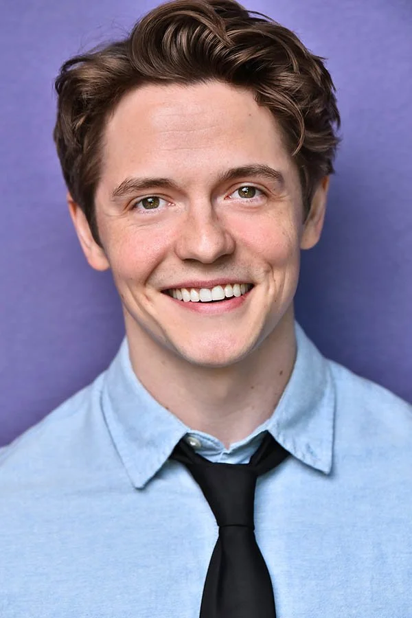 A high-impact commercial actor headshot of a man with brown hair, smiling and wearing a light blue button-down shirt with a black tie against a purple background. Directed by Marc Cartwright in Studio City to capture an authentic, approachable essenc