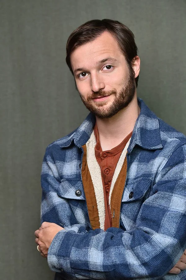 Approachable male actor headshot wearing a blue plaid jacket against a muted green background, photographed by Marc Cartwright Headshots in Los Angeles.