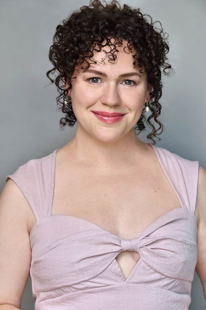 Actor with short dark curly hair smiling gently in a pale pink sleeveless top against a soft gray background.