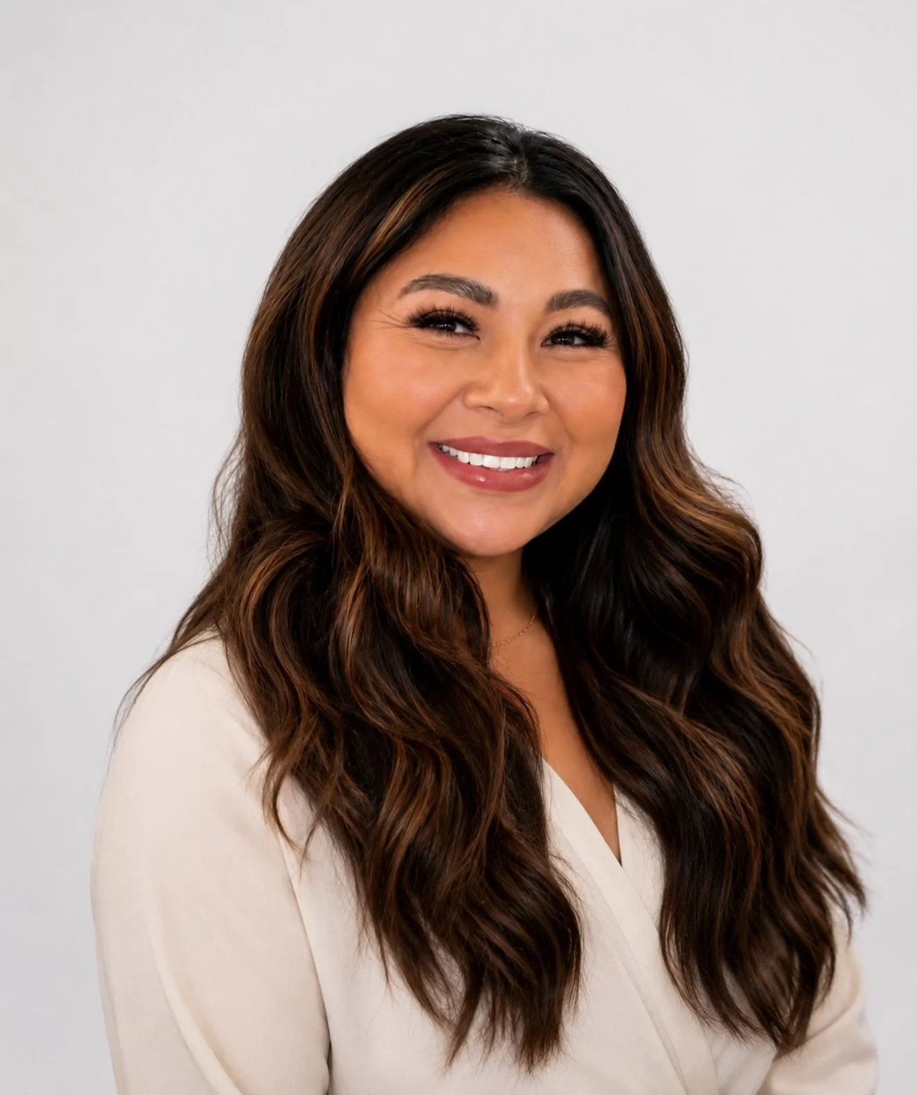 Smiling woman with long wavy brown hair wearing a white top standing against a plain background.