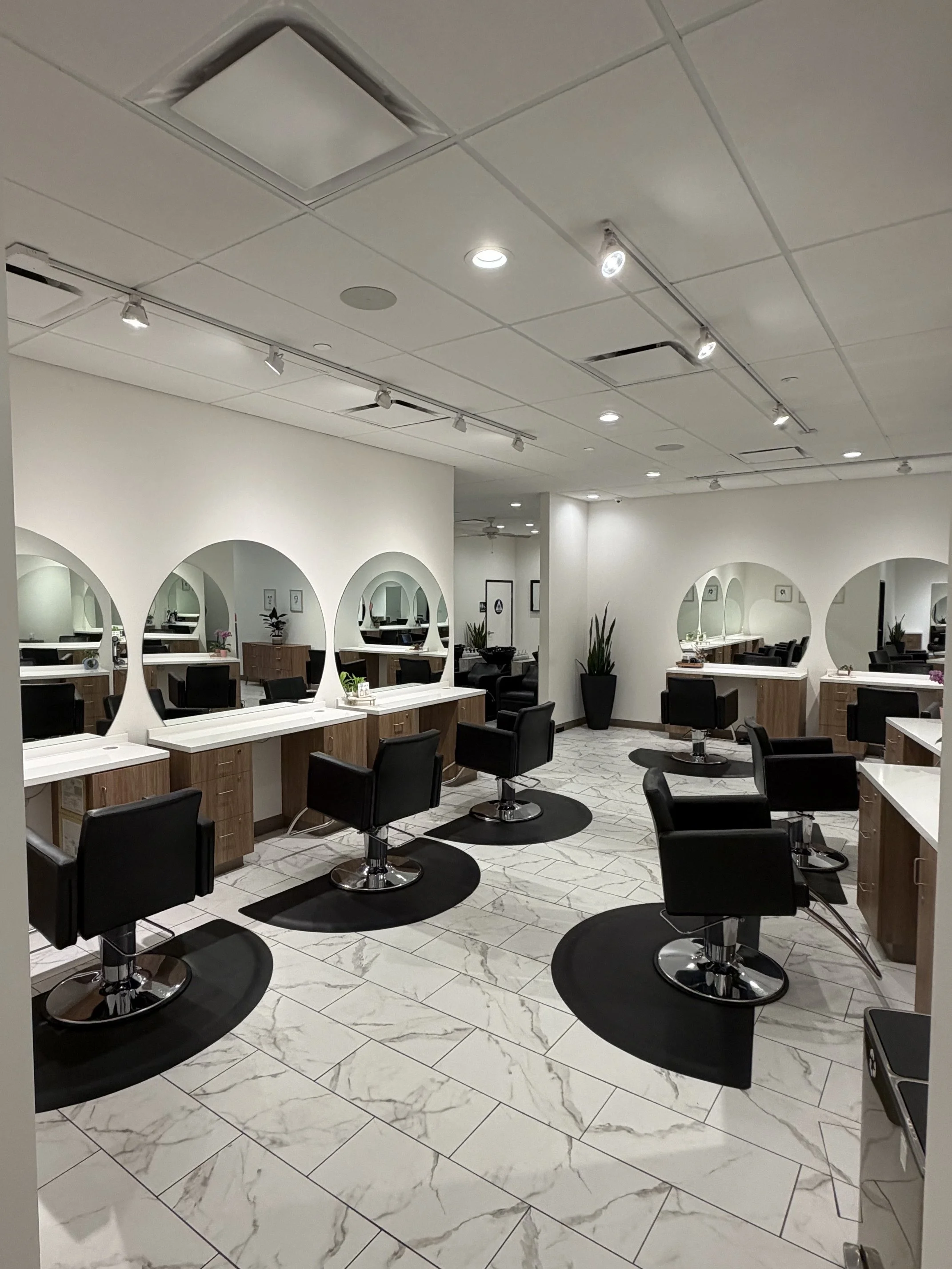 Empty hair salon with black styling chairs, white marble flooring, and multiple mirrors on the wall.