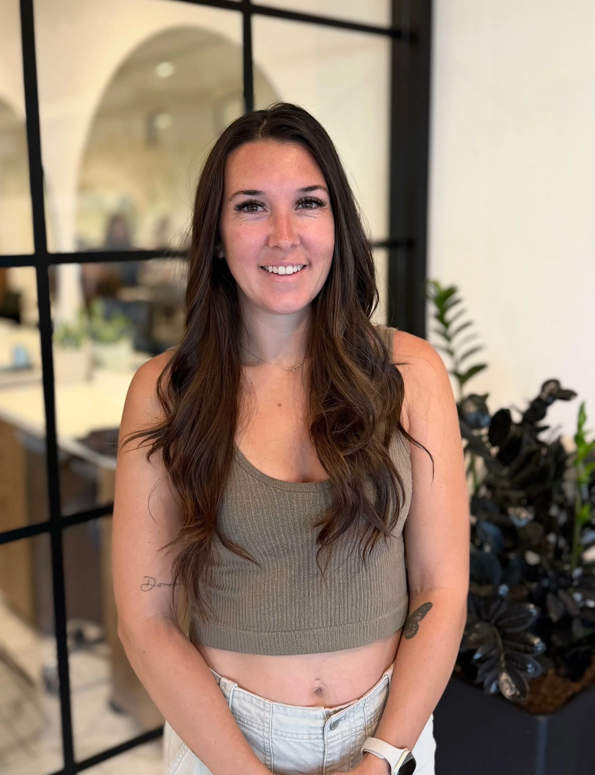 A woman with long brown hair and fair skin is smiling at the camera. She is wearing a beige ribbed tank top, beige pants, a white watch on her left wrist, and a delicate gold necklace. The background features a black grid partition, a mirror, and some potted plants.