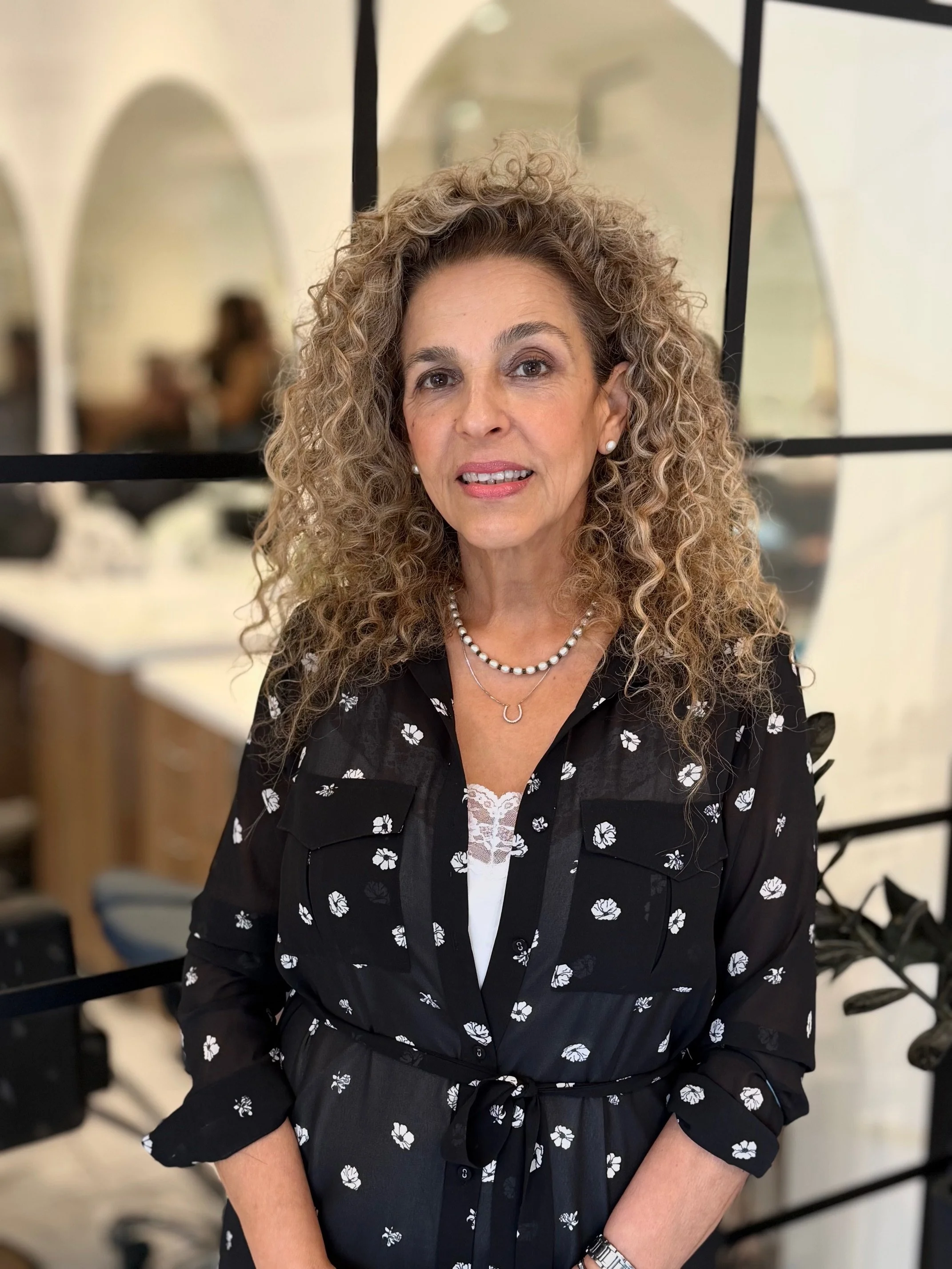 A woman with curly blonde hair and wearing pearl earrings, a pearl necklace, and a black dress with white floral patterns, sitting indoors near mirrors and black frames.