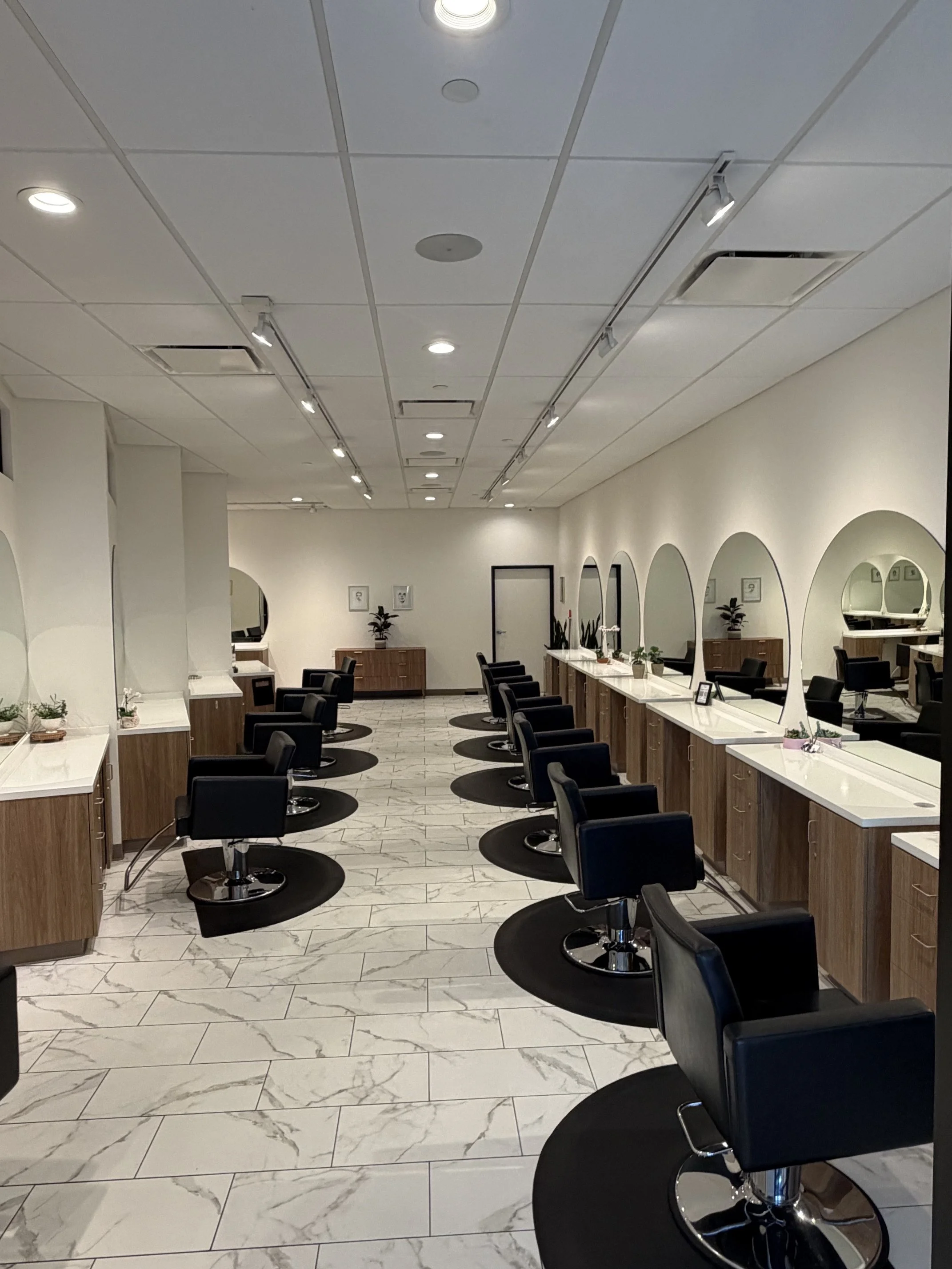 Interior of a modern hair salon with black styling chairs, white marble tile floor, and large mirrors with white frames along the wall. The ceiling has recessed lighting and track lighting, and there are small potted plants on the counters.