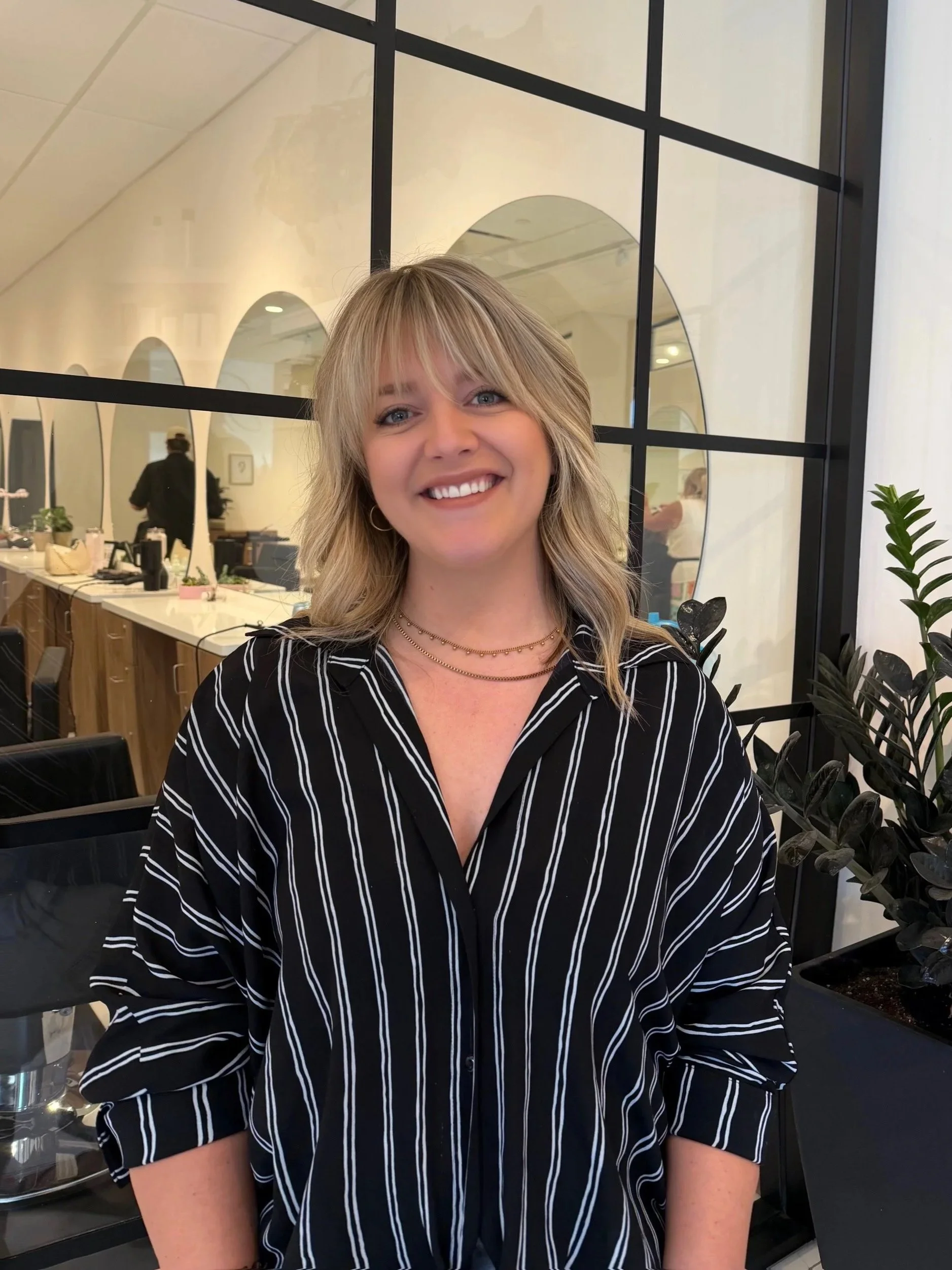 A woman with blonde hair and a striped black and white shirt smiling in a modern interior space with mirrors and plants.