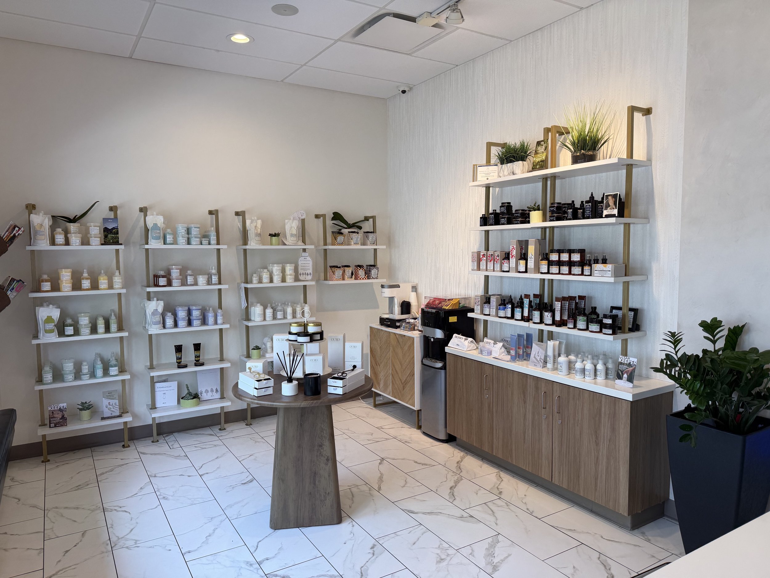 Interior of a retail shop with white shelves displaying skincare or cosmetic products, a small round wooden table with product samples, and green potted plants, on a tiled floor with a suspended ceiling.