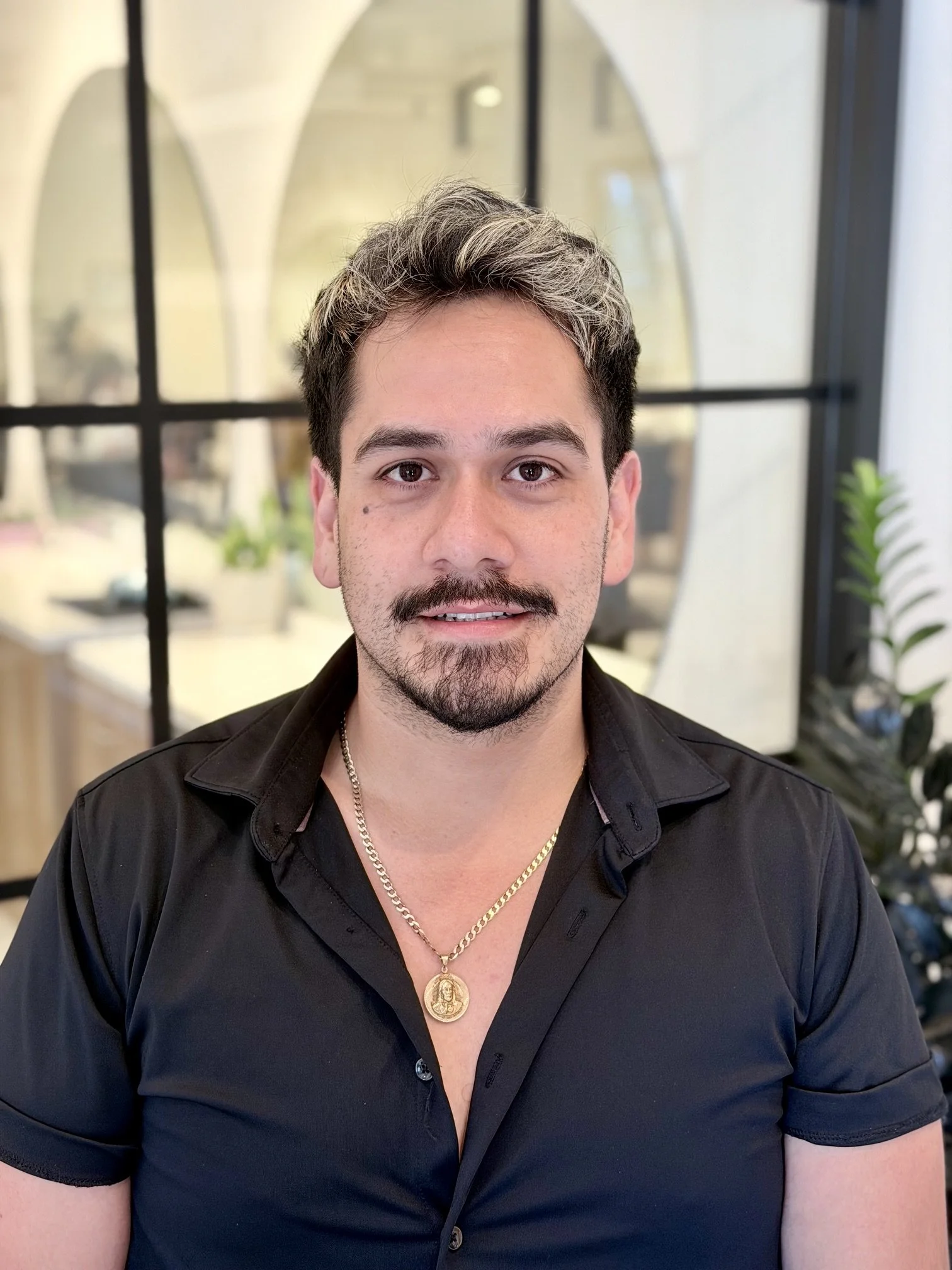 Close-up of a man with dark hair and facial hair, wearing a black shirt and gold jewelry, sitting indoors with a background of arched windows and plants.