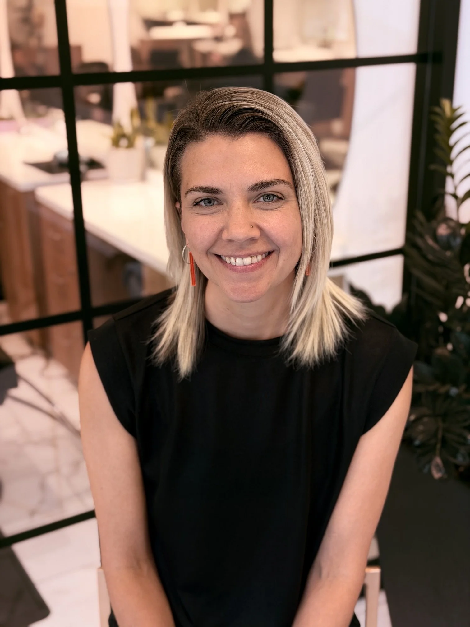 A smiling woman with blonde hair and a nose piercing, wearing a black sleeveless top, sitting in an indoor space with shelves, plants, and modern decor in the background.