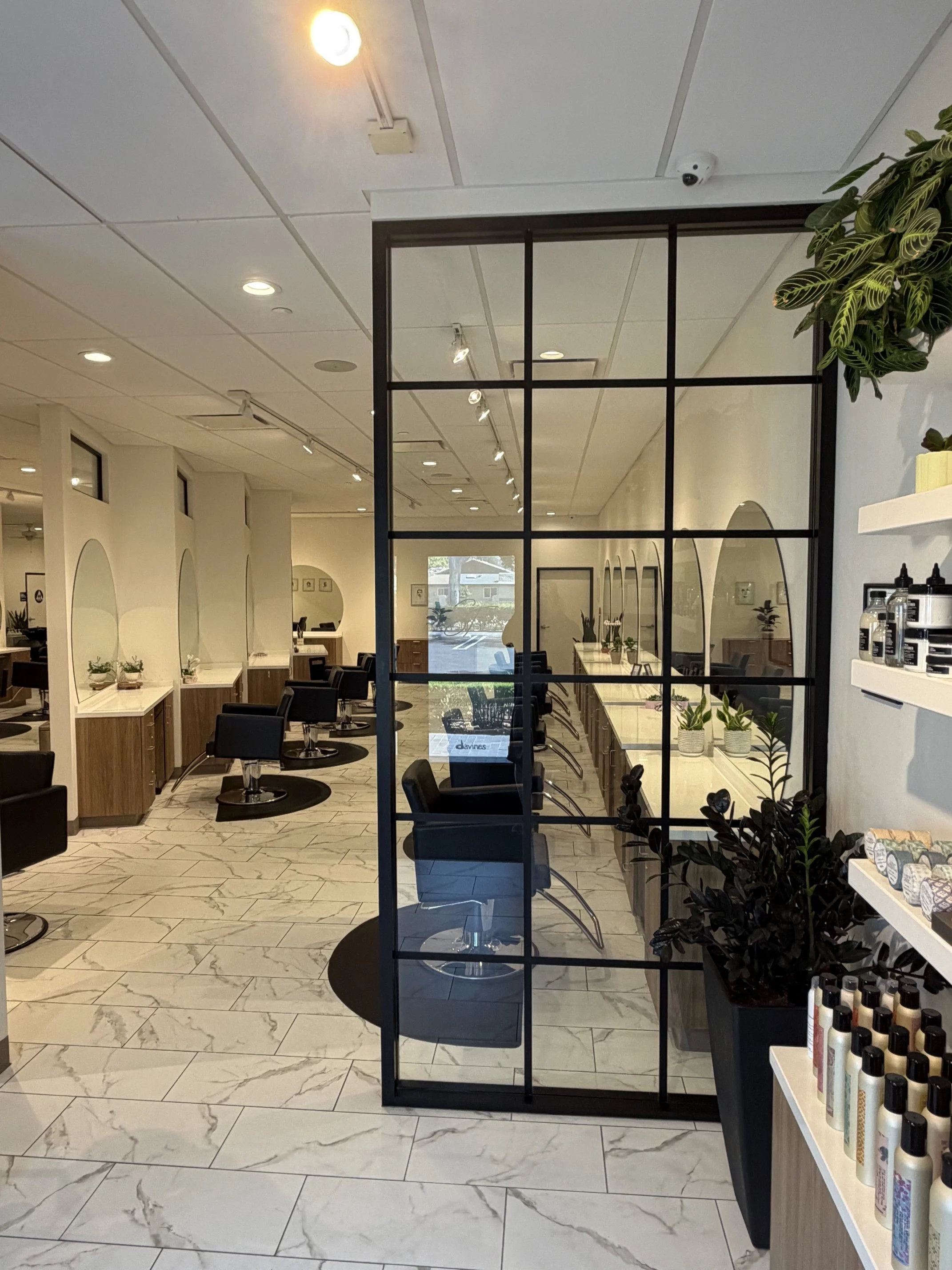Interior of a hair salon with multiple styling stations, mirrors, and chairs. Decorative plants are visible on shelves and counters, and the floor is tiled with white marble-patterned tiles.